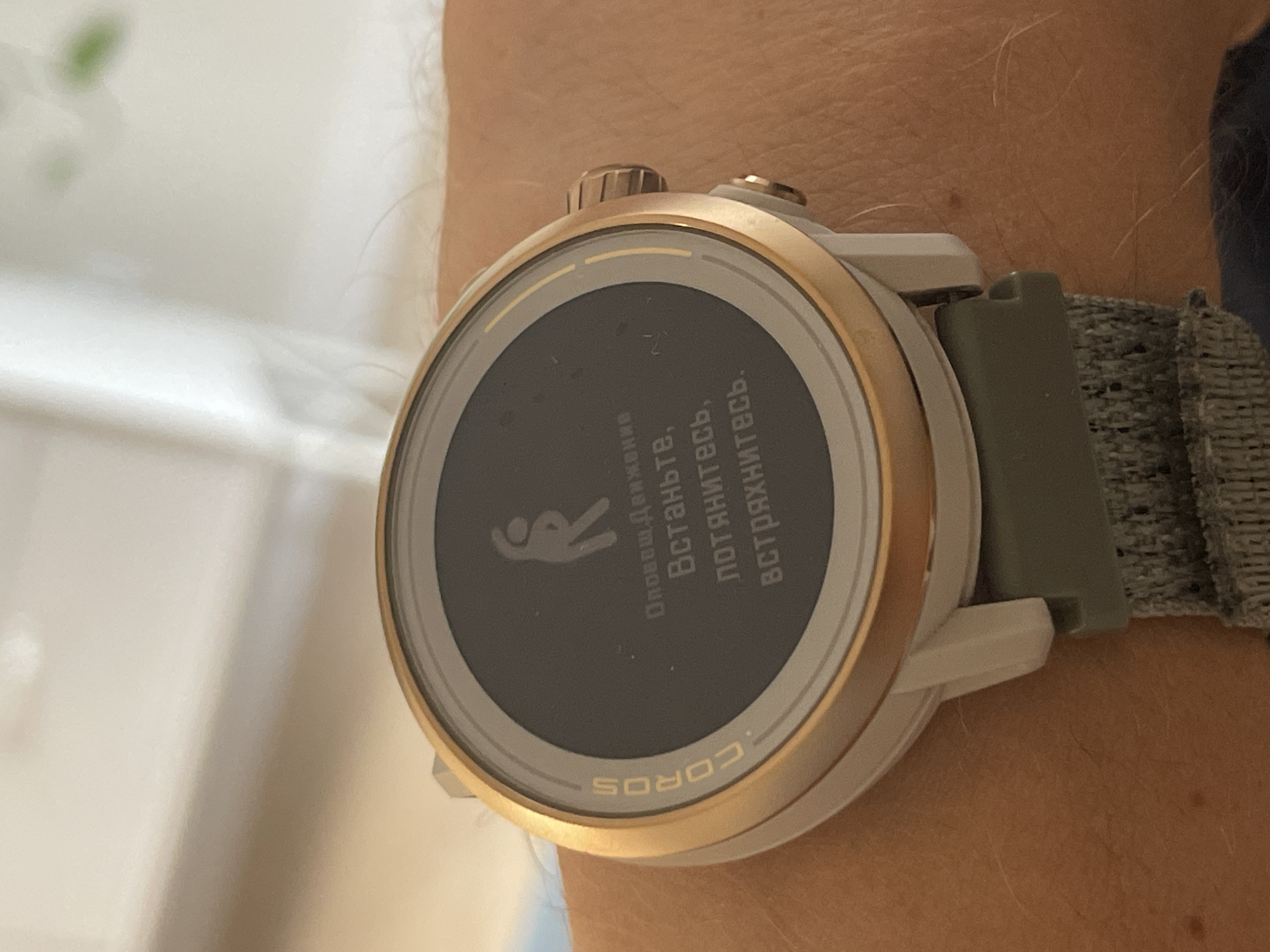 A close-up image shows a COROS brand smartwatch on a person's wrist. The watch face displays a notification in Russian, which translates to "Movement reminder. Stand up, stretch, shake". The watch has a gold-colored bezel, a light grey body, and a textured olive green watchband. The skin tone of the wrist suggests a light-skinned individual. The background is blurry and nondescript, appearing to be a light-colored wall or surface. There is no indication of the city of Izhevsk, Russia, in the image.