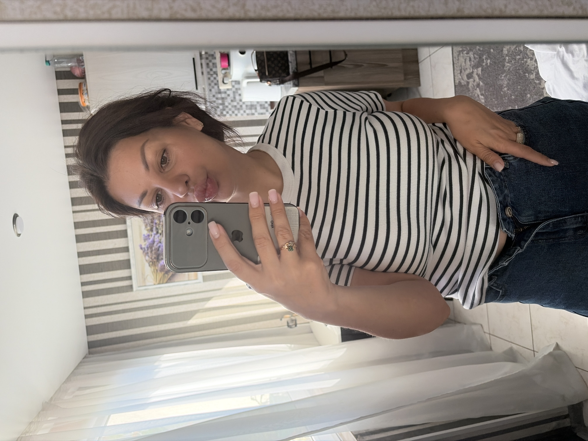 A woman with brown hair pulled back, wearing a black and white striped t-shirt and blue jeans, takes a mirror selfie in what appears to be a domestic environment. She is puckering her lips. On her left hand, she wears a ring with green stones. The background shows a window with white curtains, a striped wall, and a shelf with household items. The scene is likely indoors, possibly in an apartment in Sochi, Russia, given the context provided.