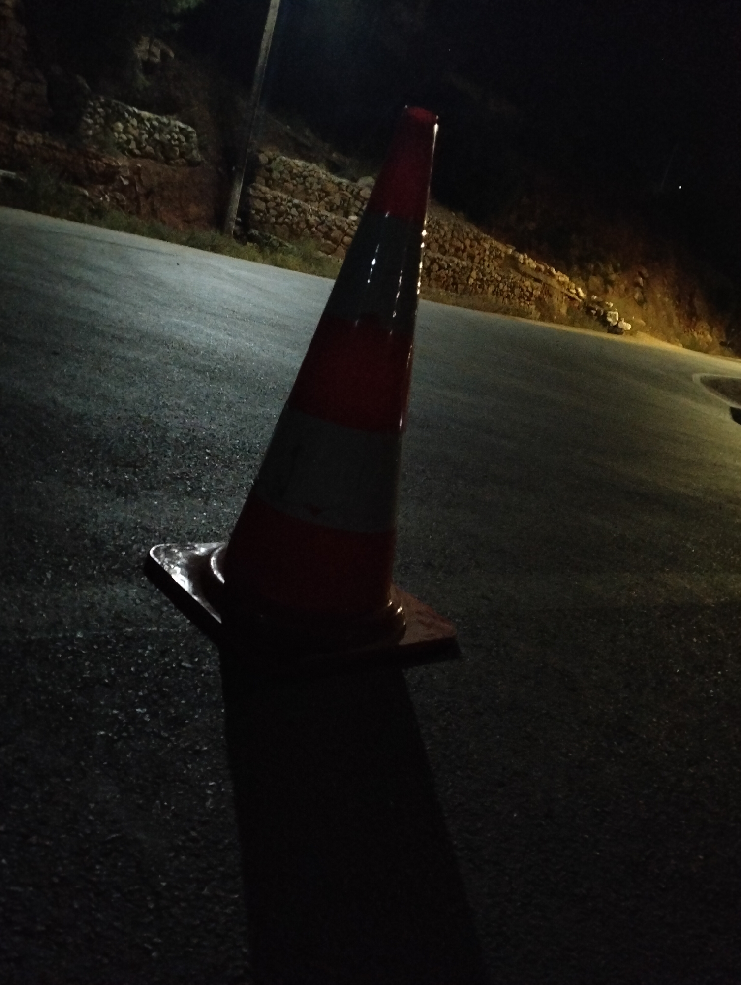A nighttime view of a red and white traffic cone on an asphalt road. The road curves into the distance. In the background, a low stone wall is visible along the roadside, suggestive of a hillside or embankment. The general location appears to be on the outskirts of Bin El Ouidane, Morocco, in an area with some elevation changes. No people or animals are visible. The lighting is dim, with the traffic cone being the primary point of light.