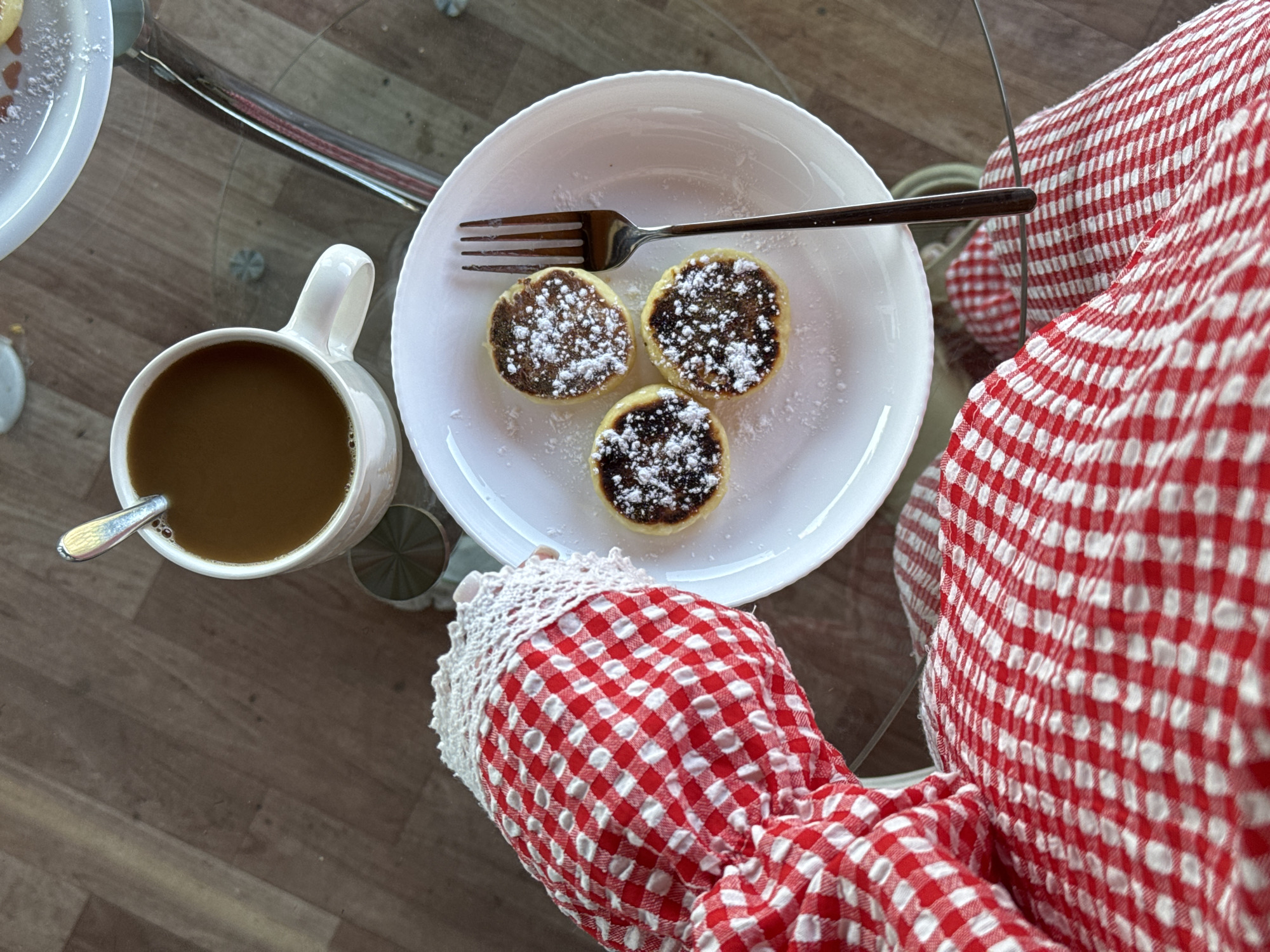 This is a top-down, eye-level shot of a breakfast scene on a glass-top table. The table is situated on a wooden floor. On the table, there is a white mug filled with coffee, a white plate holding three small, round pastries dusted with powdered sugar, and a silver fork resting on the plate. In the foreground, a person's arm and legs are visible, covered in a red and white checkered fabric. The scene appears to be indoors, likely in a home setting, and there is no indication of it being located in Minusinsk, Russia.