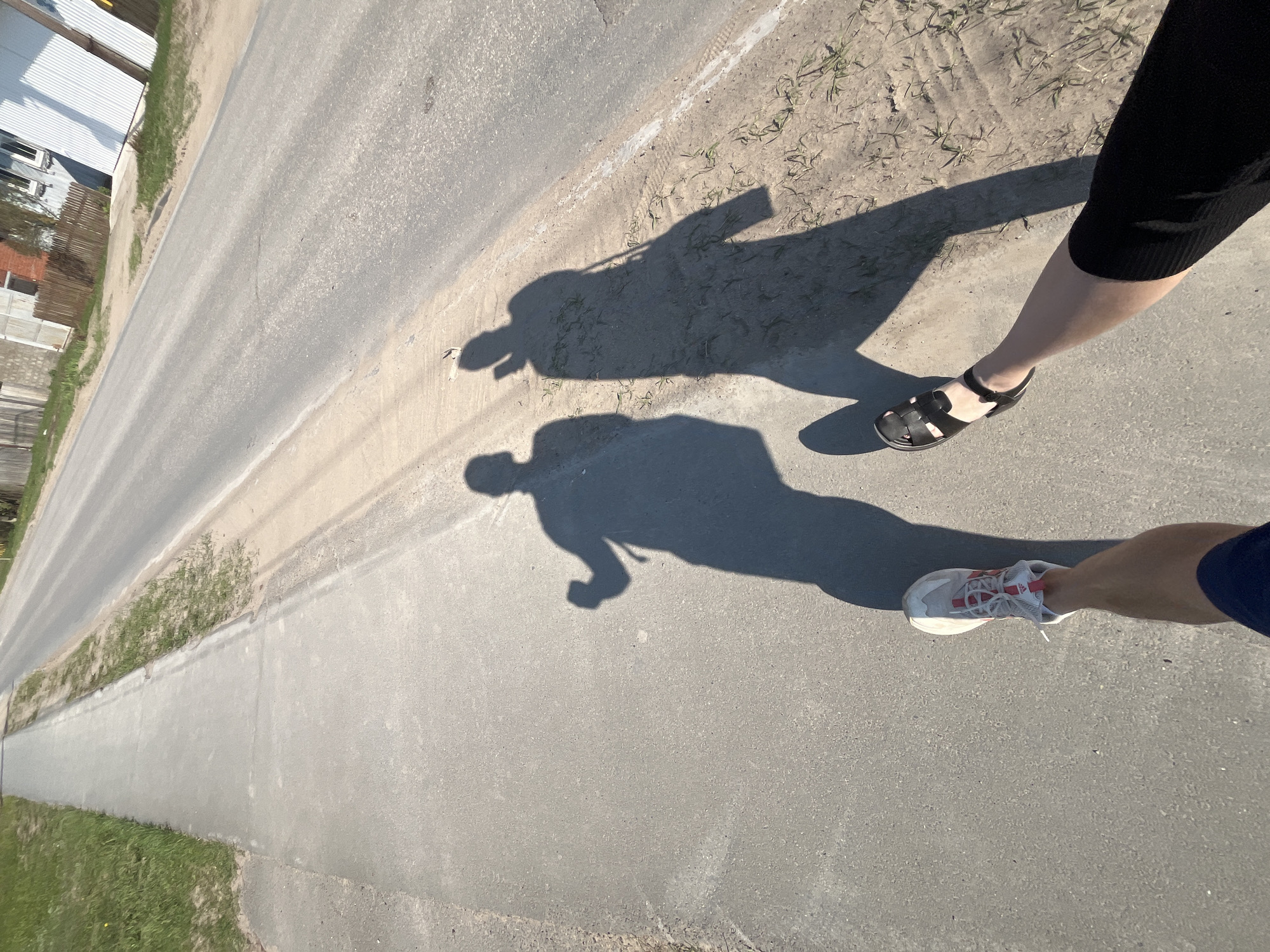 The image shows a section of paved road, likely a sidewalk, with grass and vegetation visible on one side. Two individuals are present, as indicated by their legs and shadows. One person is wearing black sandals and a black skirt, while the other is wearing white sneakers with red accents and dark blue shorts. The shadows suggest the presence of two people. It is unknown whether this location is in the city of Chaikovsky, Russia.