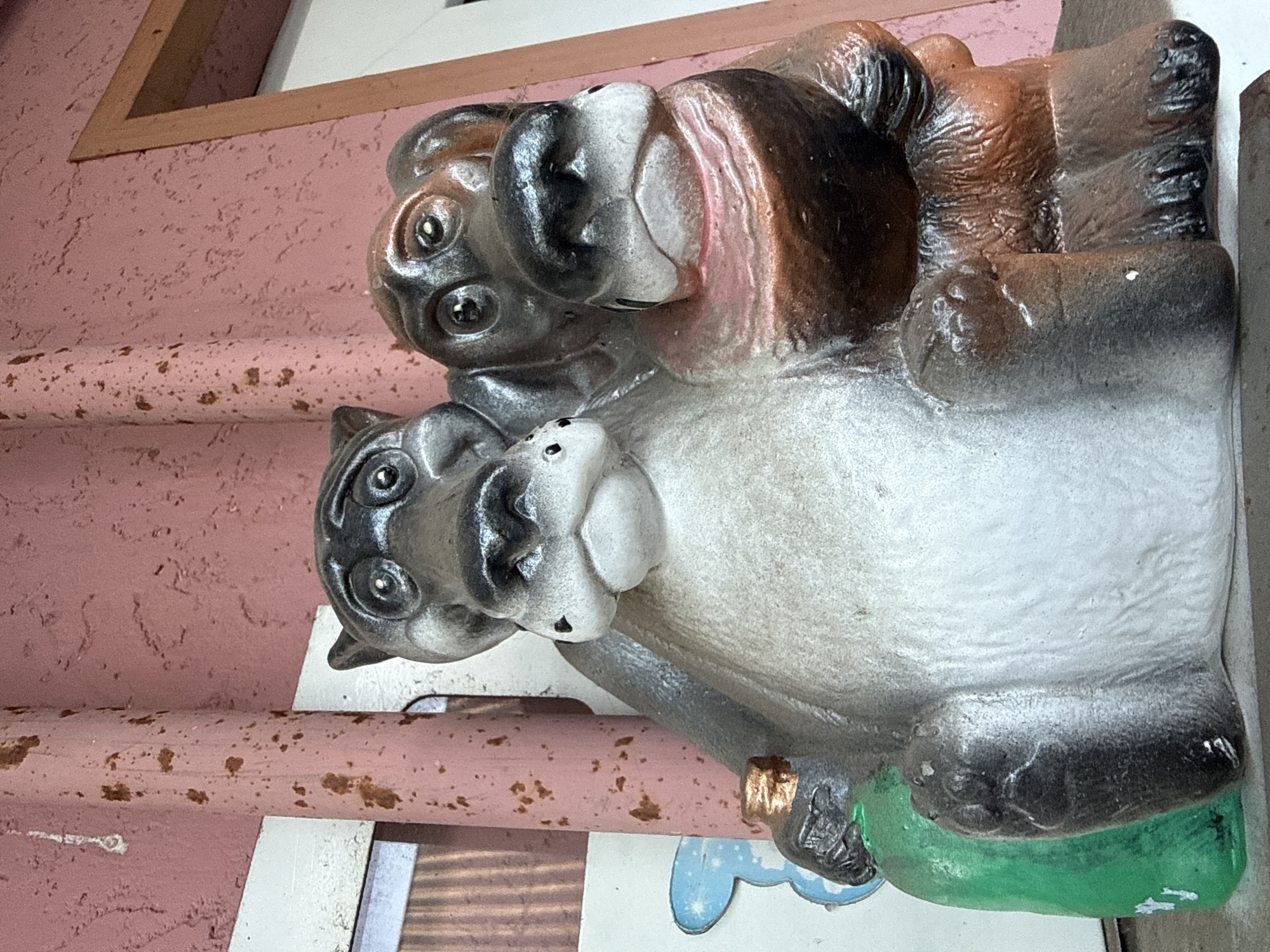 The image shows a close-up of a two-headed statue of dog-like creatures. The statue is made of a material that appears to be resin or plaster, painted in various shades of gray, brown, and white. The creature on the left has a lighter gray body and a darker gray head with large, prominent eyes and a broad snout. It is holding a green bottle with a copper-colored cap in its left arm. The creature on the right has a darker brown body and a head that is also dark brown with lighter brown accents. It has smaller eyes and a more elongated snout. The two heads are positioned very close together, as if they are sharing a single body. They are sitting down, with their legs visible in the foreground. Behind the statue, a pink, textured wall is visible, with some vertical wooden or metal posts that appear to be weathered and rusted. The overall impression is that the statue is an outdoor decoration, possibly placed in front of a building or in a yard. The location is identified as Okhtyrka, Ukraine.