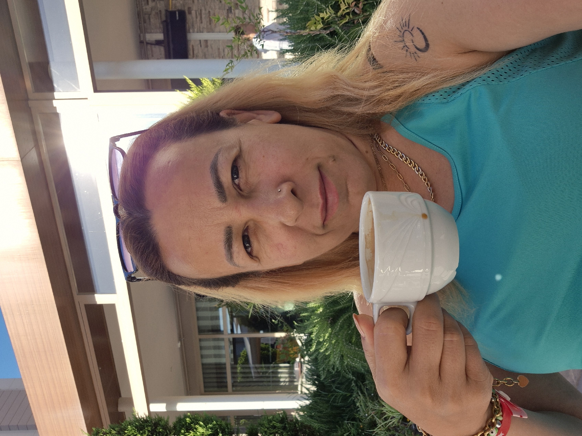 In an outdoor setting, a woman with blonde hair and light skin holds a white cup of coffee in her right hand. She is wearing a turquoise tank top, a gold chain necklace, and bracelets on her left wrist. She has a nose ring and a tattoo of a sun and moon on her right shoulder. Sunglasses are perched on top of her head. The background shows a building with a wooden overhang and a lush green garden. A man in a white shirt is visible in the distance. The lighting suggests it is daytime.