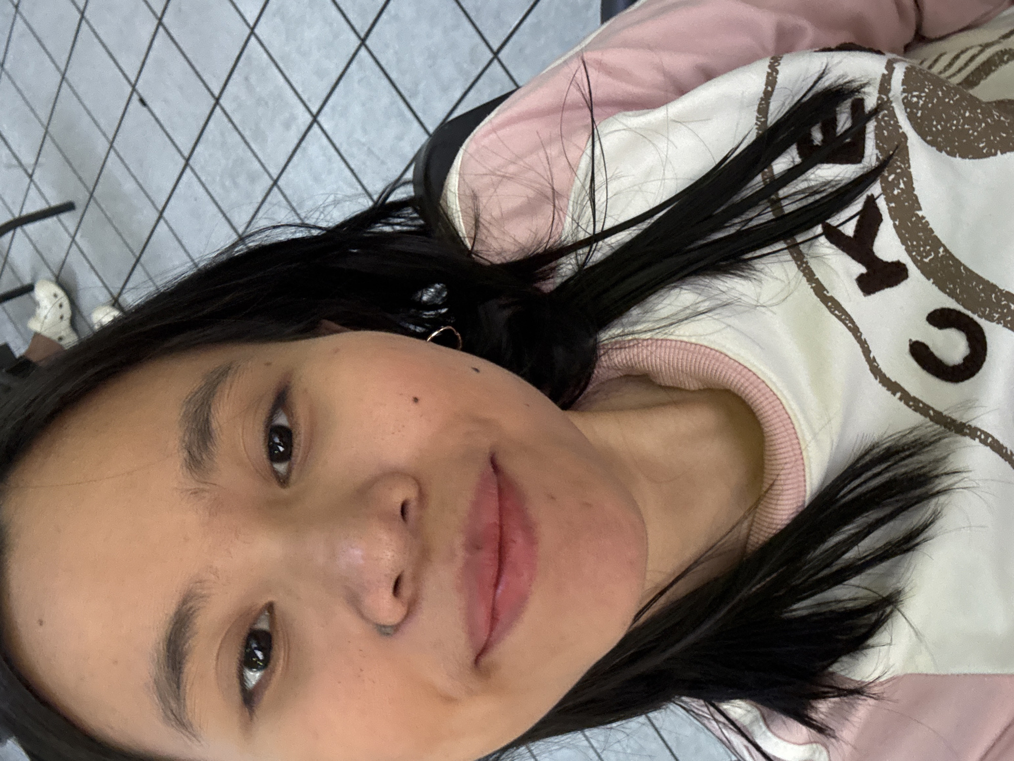 The image is a close-up selfie of a young East Asian woman in East Jakarta, Indonesia. She has long, dark hair, brown eyes, and fair skin. She is wearing a pink and white sweatshirt with brown lettering that appears to spell out "CKBE". The background is blurred, but shows a tiled floor and what looks like a table or chair. Her left ear has a small hoop earring. The woman is looking directly at the camera and smiling slightly.