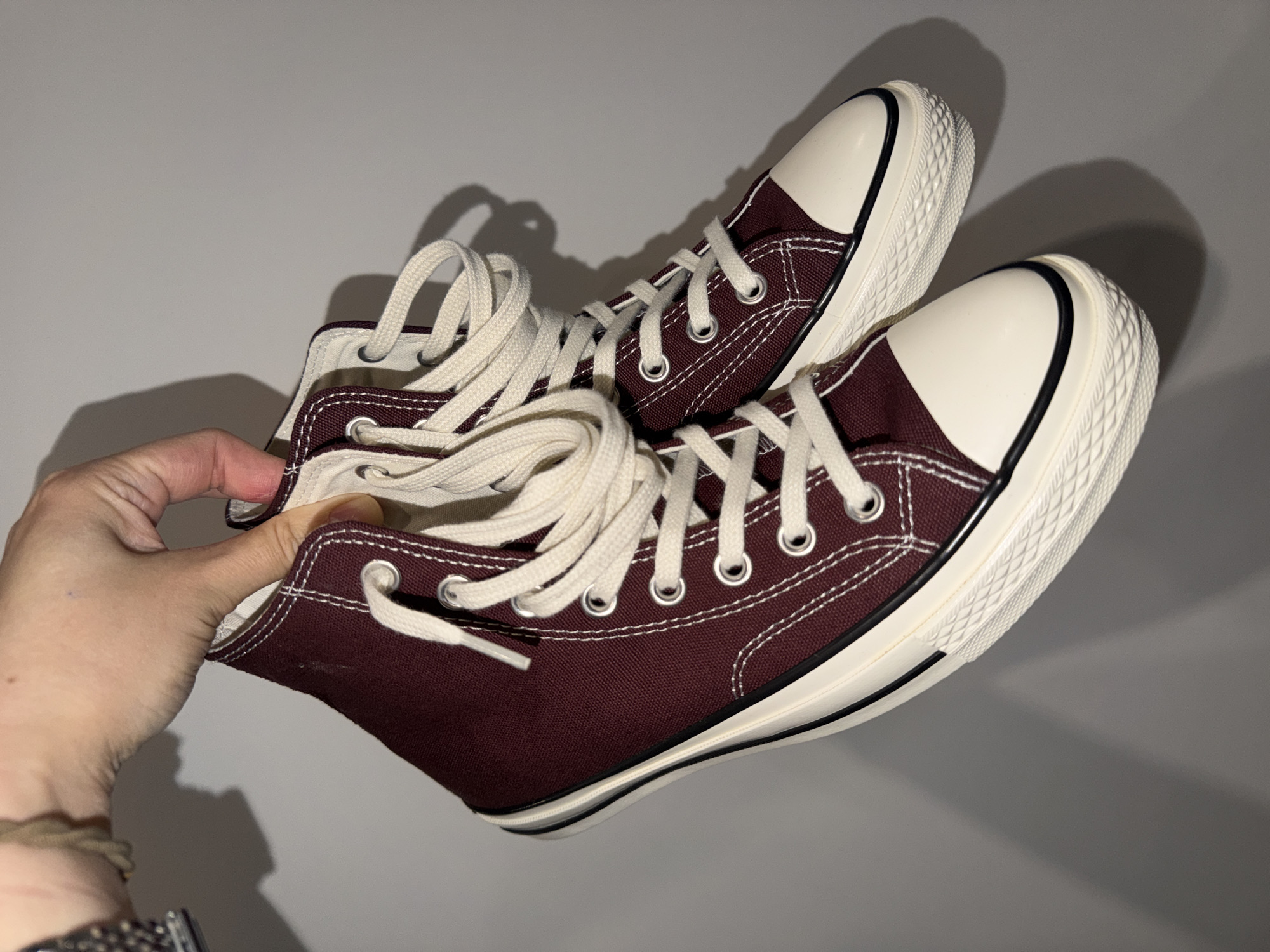 A close-up, eye-level shot against a plain light grey background shows a pair of dark maroon (burgundy) high-top canvas sneakers being held by a person's hand in Chaikovsky, Russia. The sneakers feature cream-colored laces, white contrast stitching, silver-toned metal eyelets, and cream-colored rubber toe caps and soles. A thin black stripe runs along the edge of the sole, separating it from the canvas upper. The soles have a distinct textured pattern. The laces are untied and appear somewhat loose, suggesting the shoes are new or have just been unboxed. The person's hand, with a light-colored wristband and part of a metallic bracelet visible, holds one of the sneakers near its ankle opening. Strong shadows are cast on the light background, indicating a direct light source from the side or above. No visible text is present on the shoes or in the background.