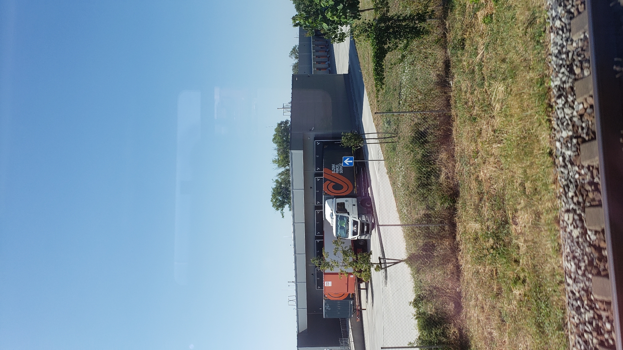 The image depicts a logistics facility on a sunny day, likely in Trnava, Slovakia. The main subject is a white MAN truck, a semi-trailer with the "SLOVAK PARCEL SERVICE" logo in orange and black, and a blue directional arrow pointing right. The truck is positioned at a loading dock of a long, modern warehouse building with a gray facade and a row of orange loading bay doors. Another trailer, orange in color and with a white "1230" label, is parked at an adjacent loading bay. A chain-link fence runs in front of the facility, separating it from a grassy area with scattered trees and bushes. In the foreground, railway tracks with ballast are visible. The sky is clear and blue.