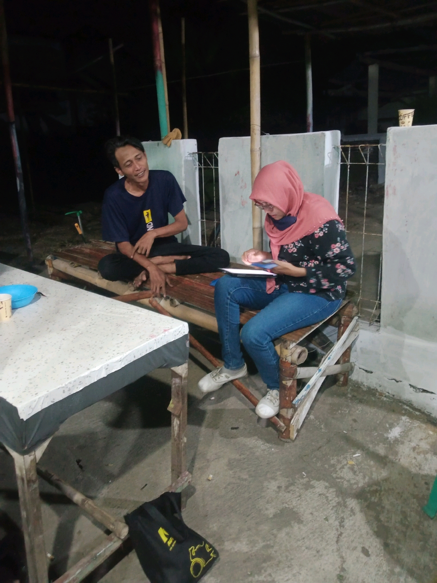 Two individuals are seated on a rustic bamboo bench outdoors at night. On the left, a young man wearing a dark blue t-shirt with a yellow emblem and black pants is looking to his right and appears to be speaking. He is sitting cross-legged. On the right, a young woman, wearing a pink headscarf, glasses, a floral patterned long-sleeved shirt, and blue jeans, is looking down at a tablet she holds in her hands, with her fingers interacting with the screen. She is wearing white sneakers. In the foreground, a white-topped table with a dark tablecloth is positioned to the left, with a blue bowl and a paper cup on it. Behind the people, a white wall with a metal fence and bamboo poles are visible, suggesting an open-air structure. The ground is concrete. A black bag with a yellow design is on the ground near the table. The scene is illuminated by artificial light, casting shadows and creating a dim atmosphere. This appears to be a casual gathering or relaxation spot in Rejasari, Indonesia.