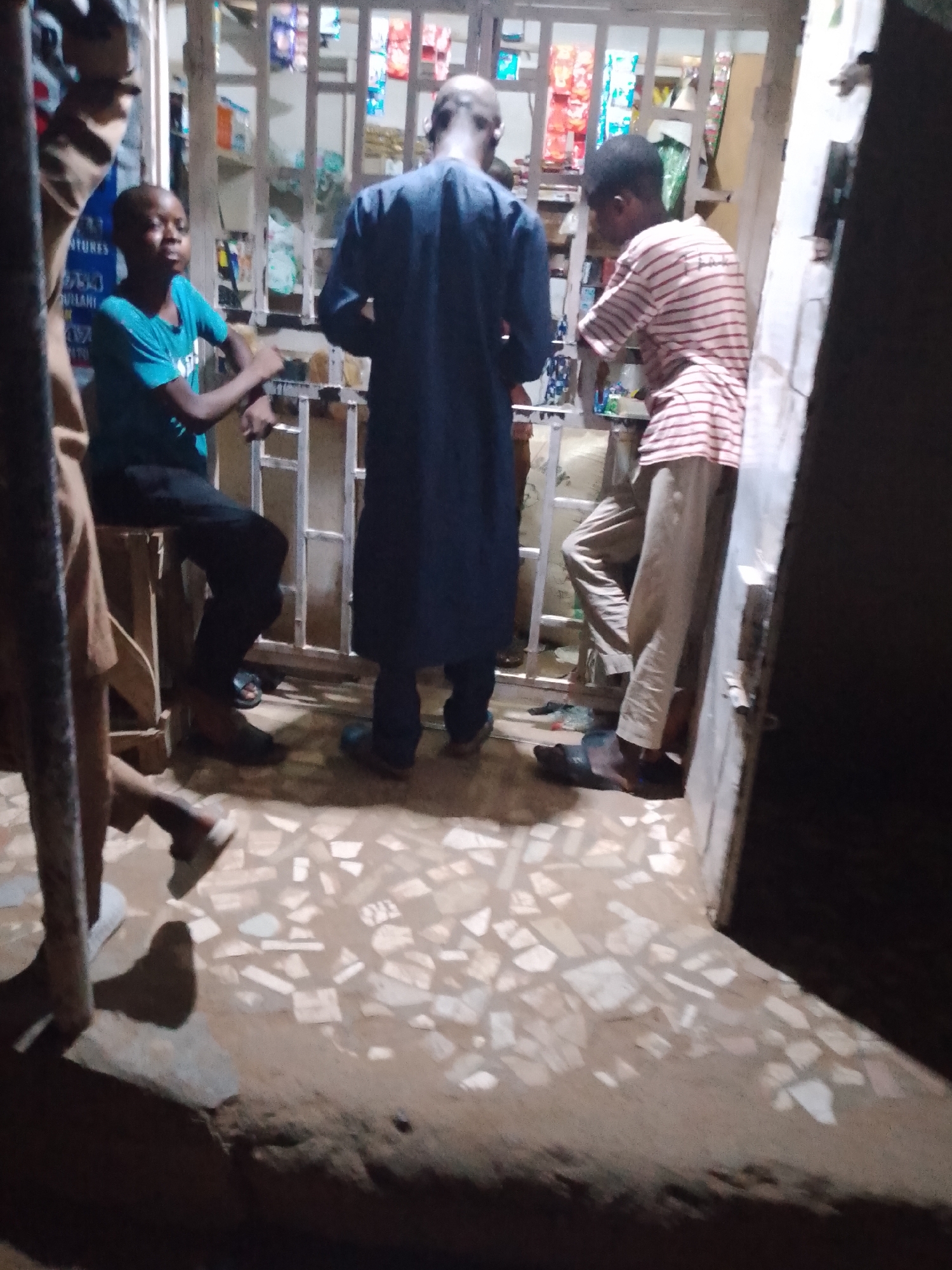 In Arawa, Nigeria, three people are visible inside a small store. One individual, wearing a blue shirt and black pants, is seated. Another, clad in a long navy garment, stands with their back facing the viewer. The third person, in a horizontally striped shirt and light-colored pants, leans against the entrance. The store interior displays an array of packaged goods behind a metal grate. The floor has a light mosaic tile pattern. The scene is dimly lit.