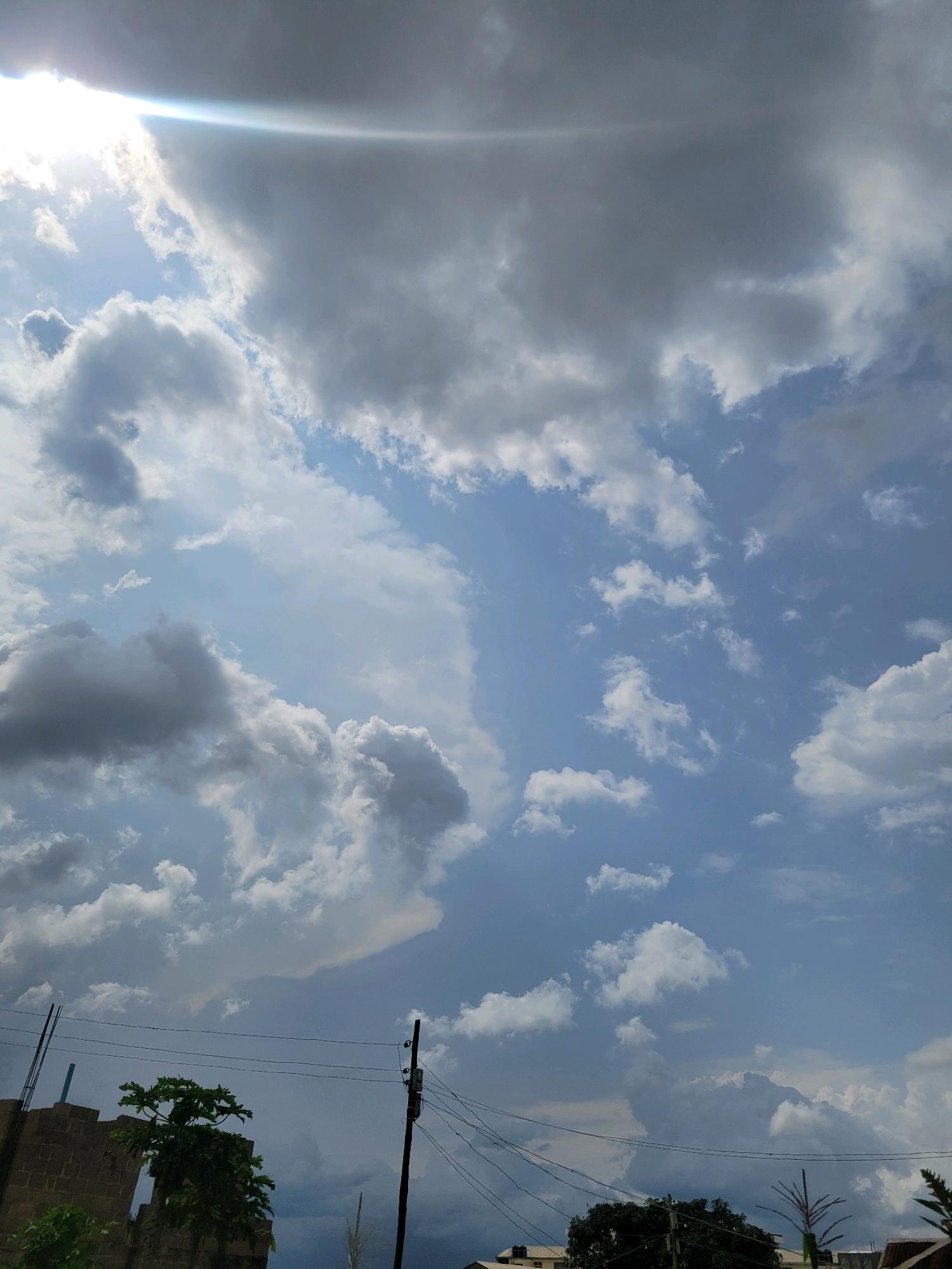 This is a landscape photograph taken in the city of Jos, Nigeria. The image predominantly features a dramatic sky filled with a mix of dark, ominous clouds and brighter, wispy formations. Sunlight pierces through the clouds in the upper left quadrant, creating a lens flare effect. Below the sky, a line of utility poles and wires stretches across the lower portion of the frame. To the left, a partially constructed brick building is visible, with a lush green tree growing in front of it. Further to the right, another utility pole with complex wiring is prominent. The overall scene suggests a potentially stormy or post-storm environment in an urban or semi-urban area of Jos.