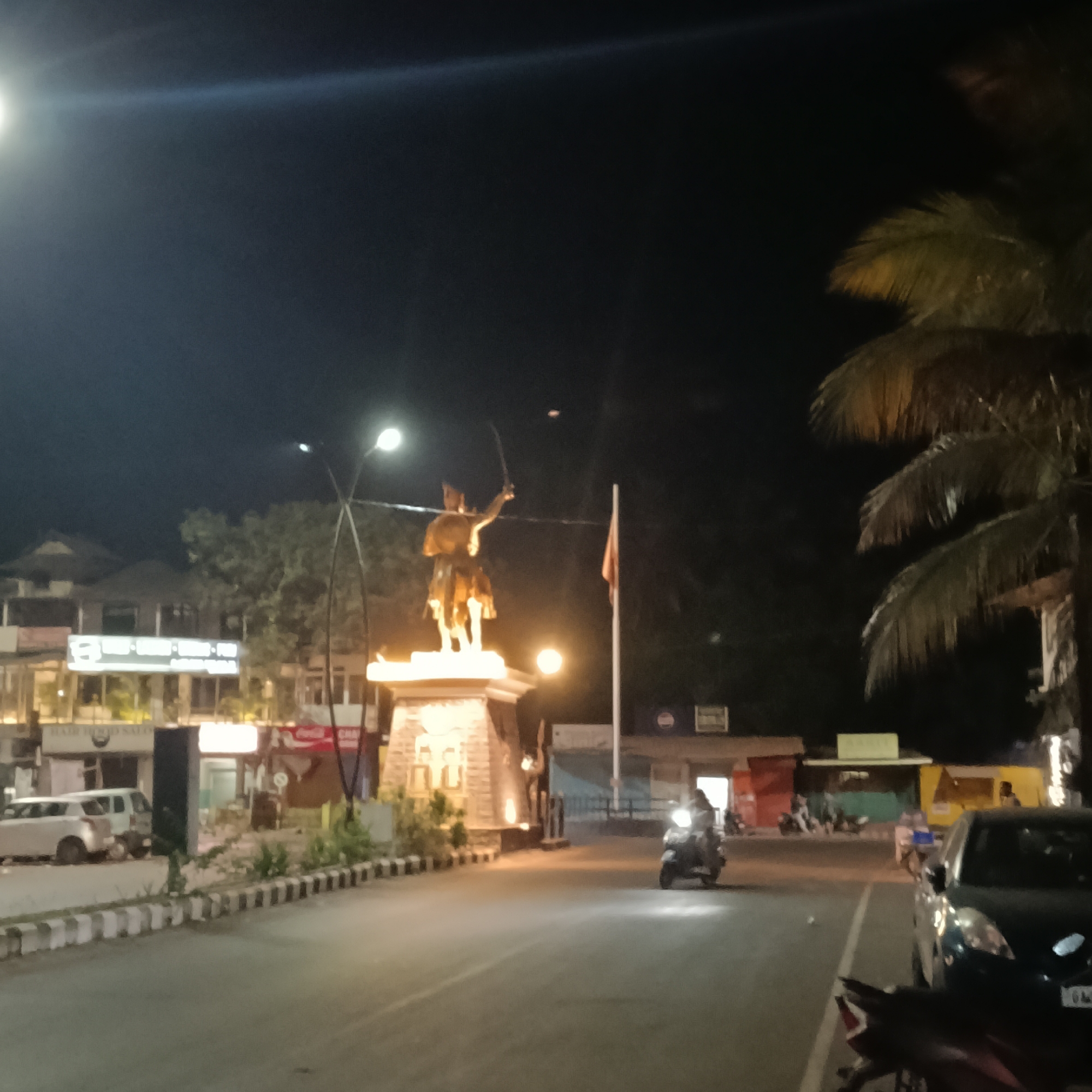 The image depicts a night scene in what appears to be a city or town square in Oxel, India. In the center is a lit statue on a stone pedestal, likely representing a historical figure, holding a sword, in an open square. A flag is also visible nearby. A motorcycle is on the road approaching the statue. Palm trees are present on the right side. Buildings line the road, appearing to be shops or commercial establishments. Several parked vehicles, including a white car and a darker colored car, are on the side of the road. Pedestrians are faintly visible near the buildings. Streetlights illuminate the scene. The general environment suggests a well-lit but relatively quiet night.