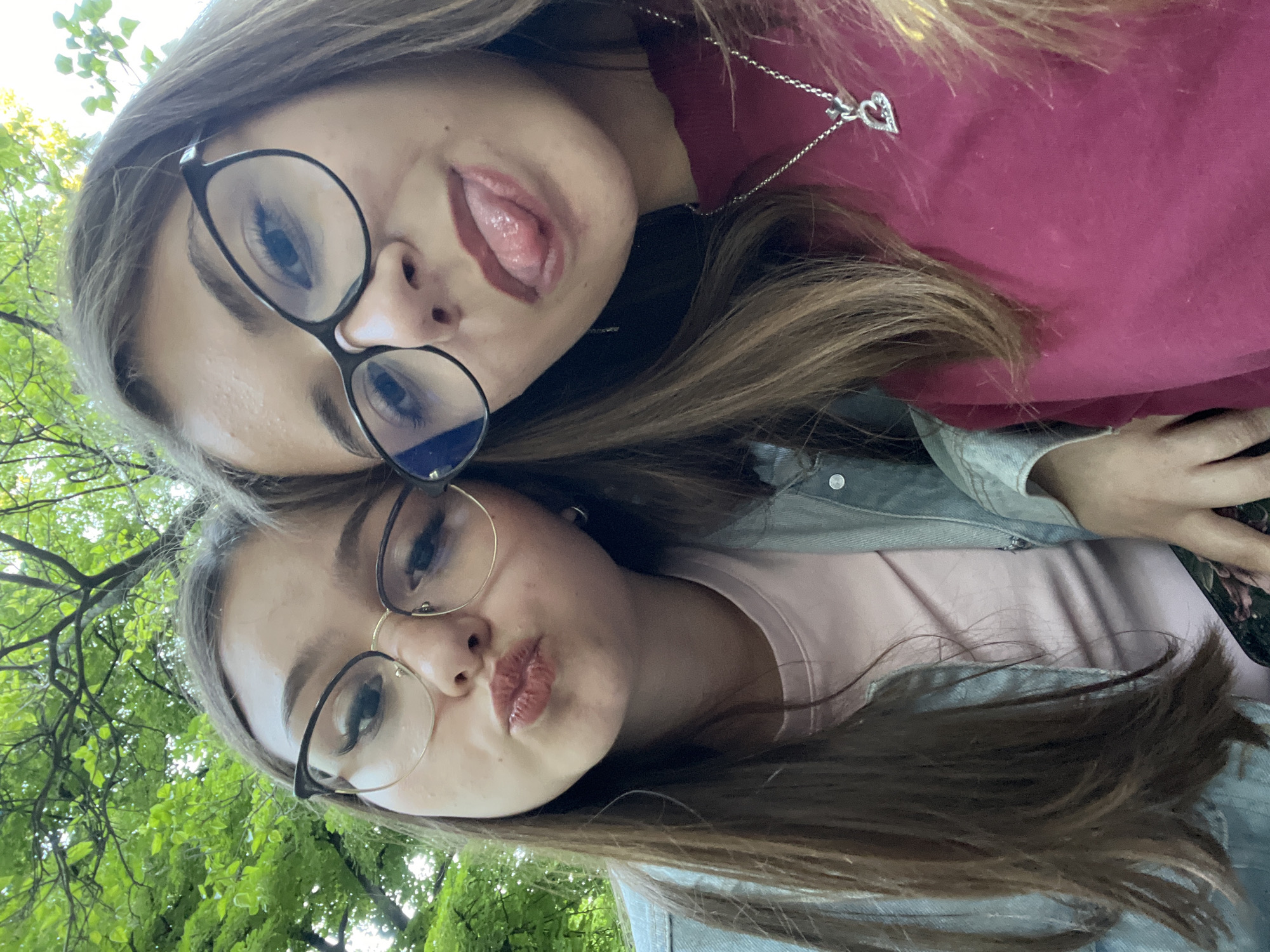 Two young women are taking a selfie outdoors. The woman on the top is sticking her tongue out and wearing glasses, a maroon sweater, and a heart necklace. The woman on the bottom is making a kissing face and wearing glasses and a light pink shirt with a denim jacket. Green foliage is visible in the background. The location is most likely within a park or garden in Plovdiv, Bulgaria, given the presence of lush greenery.