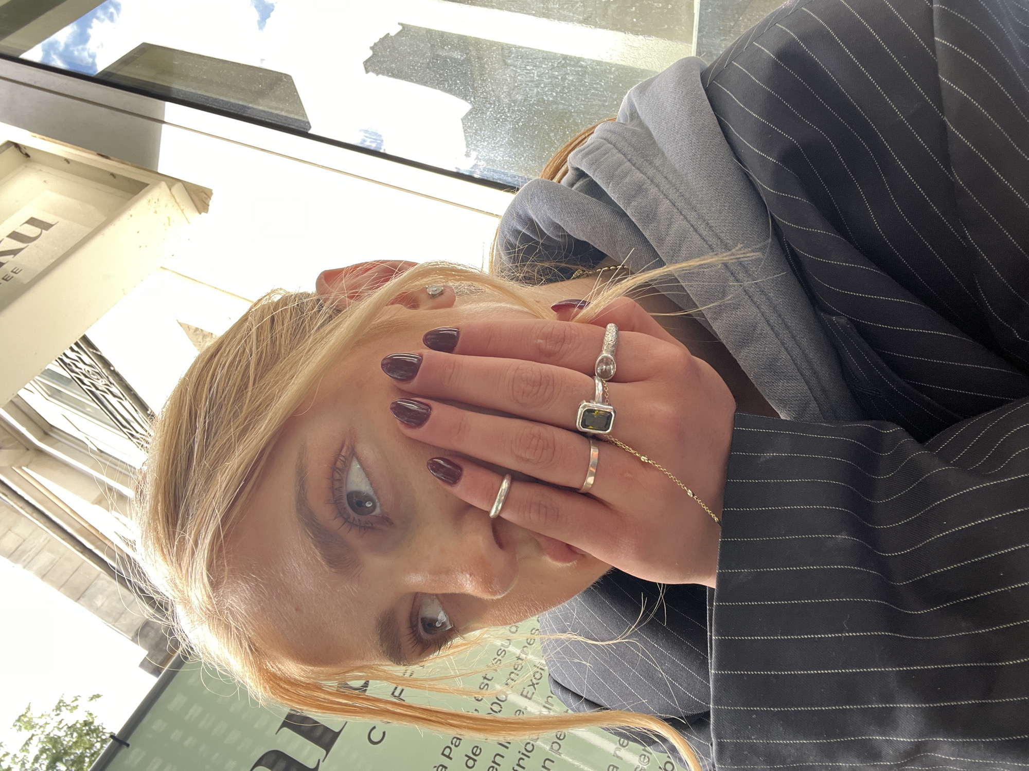 A young woman with blonde hair, dark red nail polish, and multiple rings on her fingers poses at a low angle, partially obscuring her face with her hand. She is wearing a gray hoodie under a pinstripe jacket. The background includes a window with reflections and a sign that reads "à Paris de... en... est issu a... 100 mètres... Exci...un pe...a re..." which suggests she is in Paris, France.