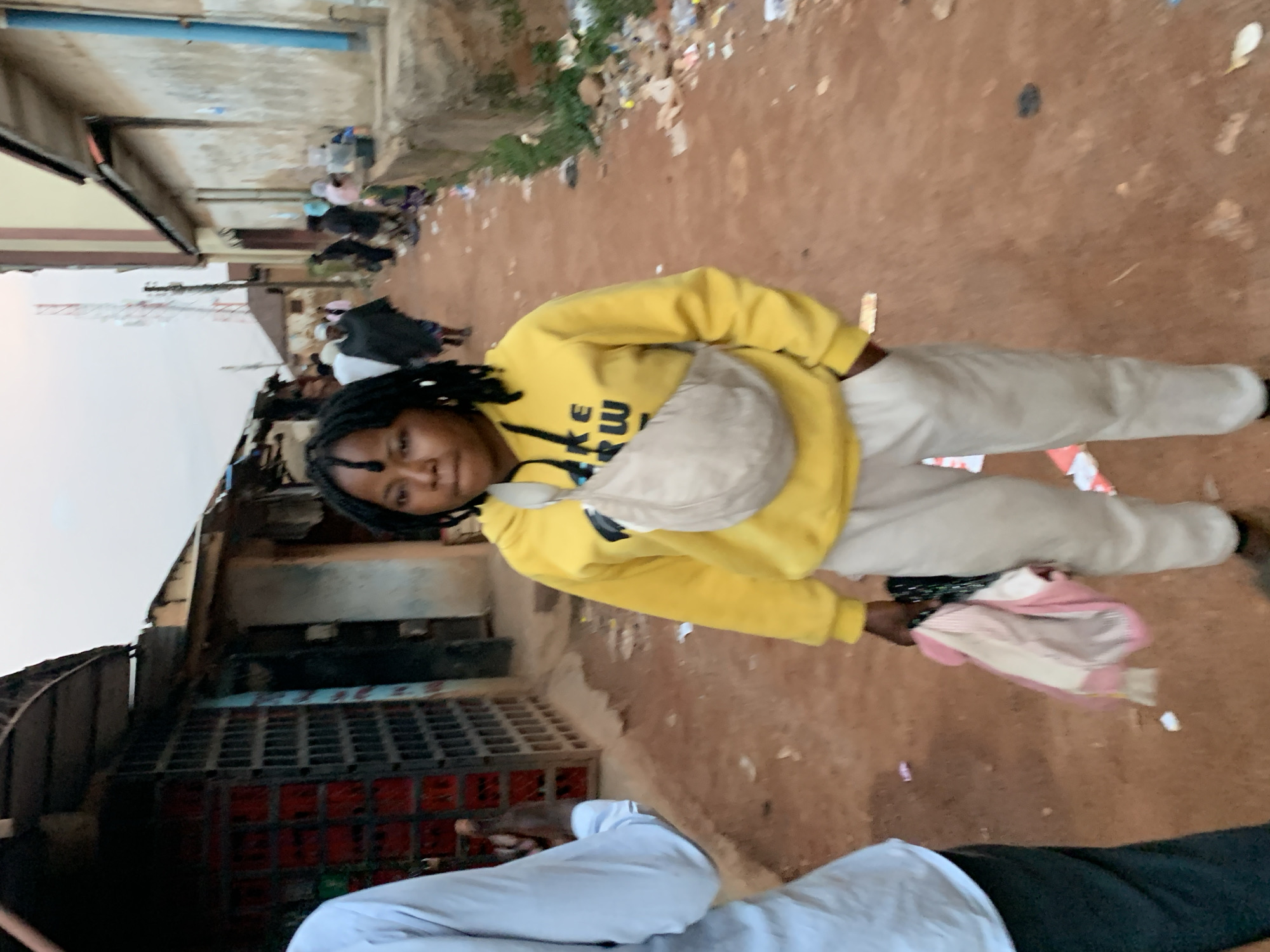 In the city of Jos, Nigeria, a young Black woman with dreadlocks walks towards the camera. She is wearing a yellow hooded sweatshirt with the partial text "KE RW" visible, and light-colored sweatpants. A beige fanny pack is worn across her chest, and she is holding a bundle of pink and white fabric in her right hand. A person wearing a light blue shirt and dark pants is partially visible on the left, walking away from the camera. The background depicts a dusty, reddish-brown dirt road lined with low-rise buildings and market stalls. Debris, including plastic bags and wrappers, is scattered along the roadside. In the distance, other individuals are seen walking and milling about, and a telecommunications tower is visible against an overcast sky. The scene suggests a bustling urban environment, likely a market area or residential street in Jos.
