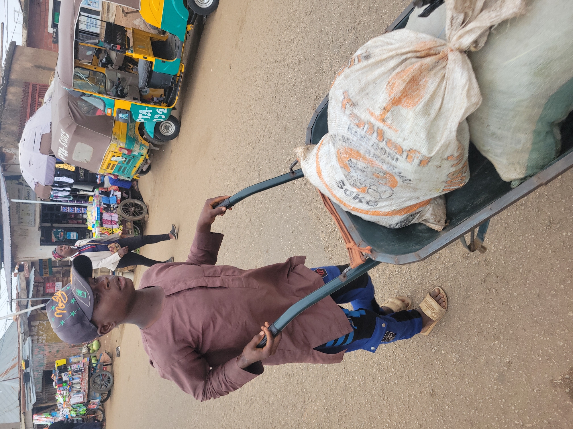 In Jos, Nigeria, a young boy in a brown tunic and blue pants with sandals pushes a wheelbarrow filled with two large sacks on a dusty, unpaved street. The boy wears a dark baseball cap with green lettering and designs. In the background, several market stalls are visible, with various goods displayed, including items on carts and shelves. A yellow and teal colored auto rickshaw is parked on the right side of the image. A woman in a white jacket and headscarf is walking away from the camera. The sky is overcast.