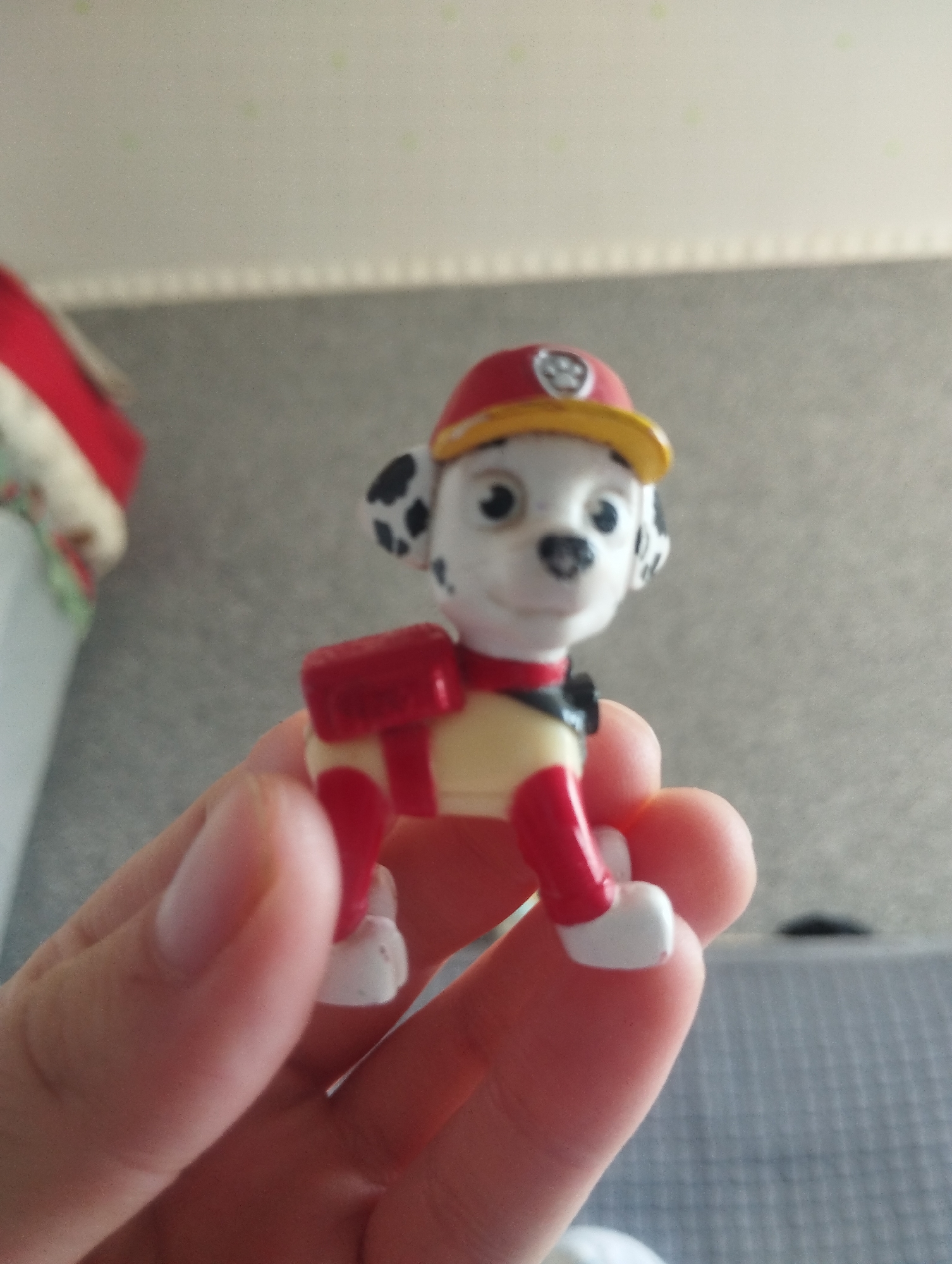 A close-up, eye-level, indoor shot shows a hand holding a small, plastic toy dog character. The toy dog is white with black spots on its ears and has a red and yellow firefighter's helmet. It is wearing a yellow shirt with red sleeves and a red backpack. The background is out of focus and appears to be a wall with some pattern and a blurred object on the left. The lighting suggests an indoor setting. The location is Orlovka, Kyrgyzstan.