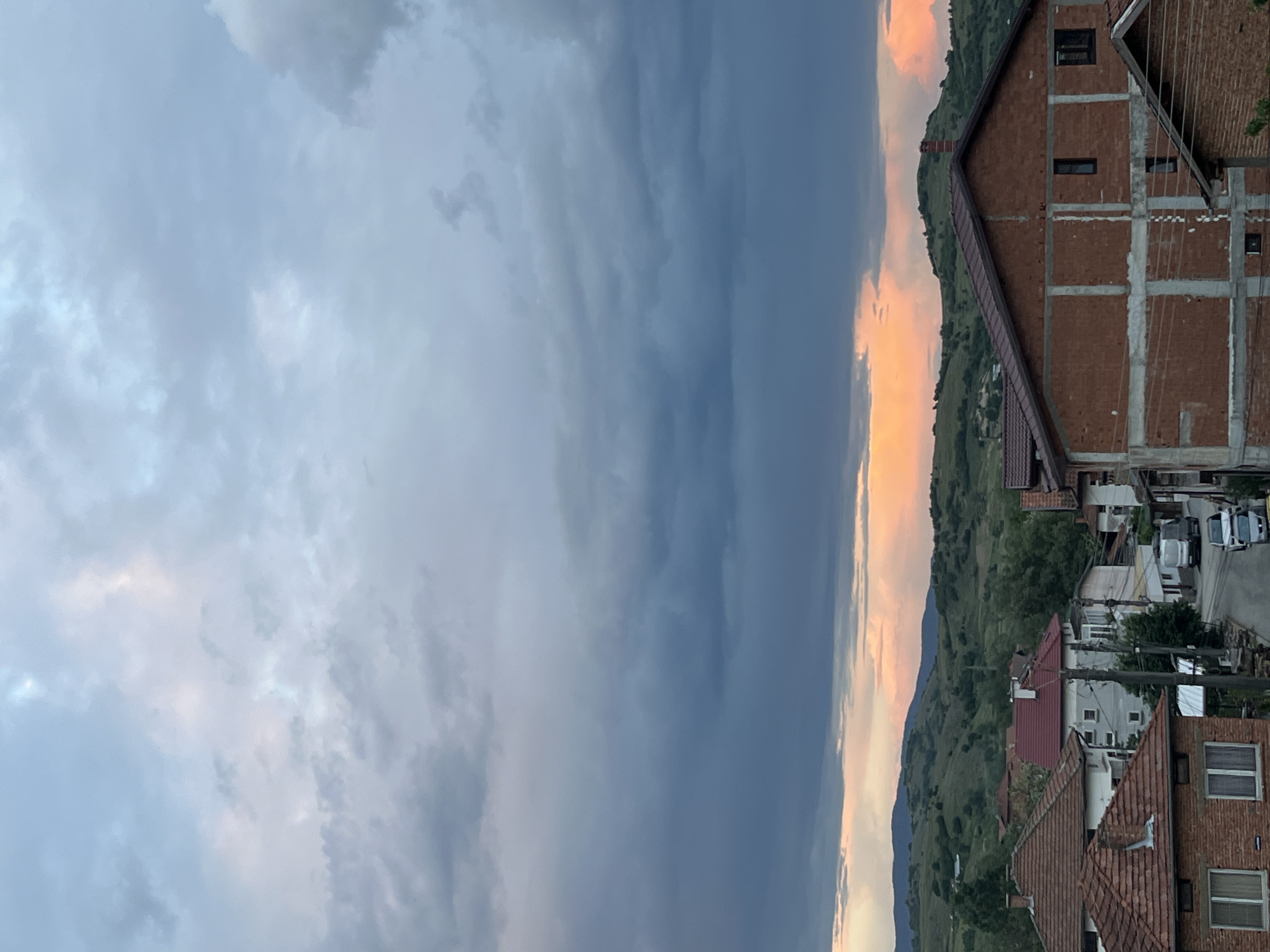 The image shows a view of Kornitsa, Bulgaria. The sky occupies the left side of the image, with varying shades of grey clouds. On the right side, a mountain range is visible, illuminated by a setting sun that casts a warm orange glow along the horizon. A residential area lies along the base of the mountain range. Brick houses with tiled roofs are prominent, and a larger, unfinished brick building is noticeable with its concrete supports exposed. A street runs through the residential area, with several parked cars visible. Greenery dots the landscape. The orientation of the image is sideways.