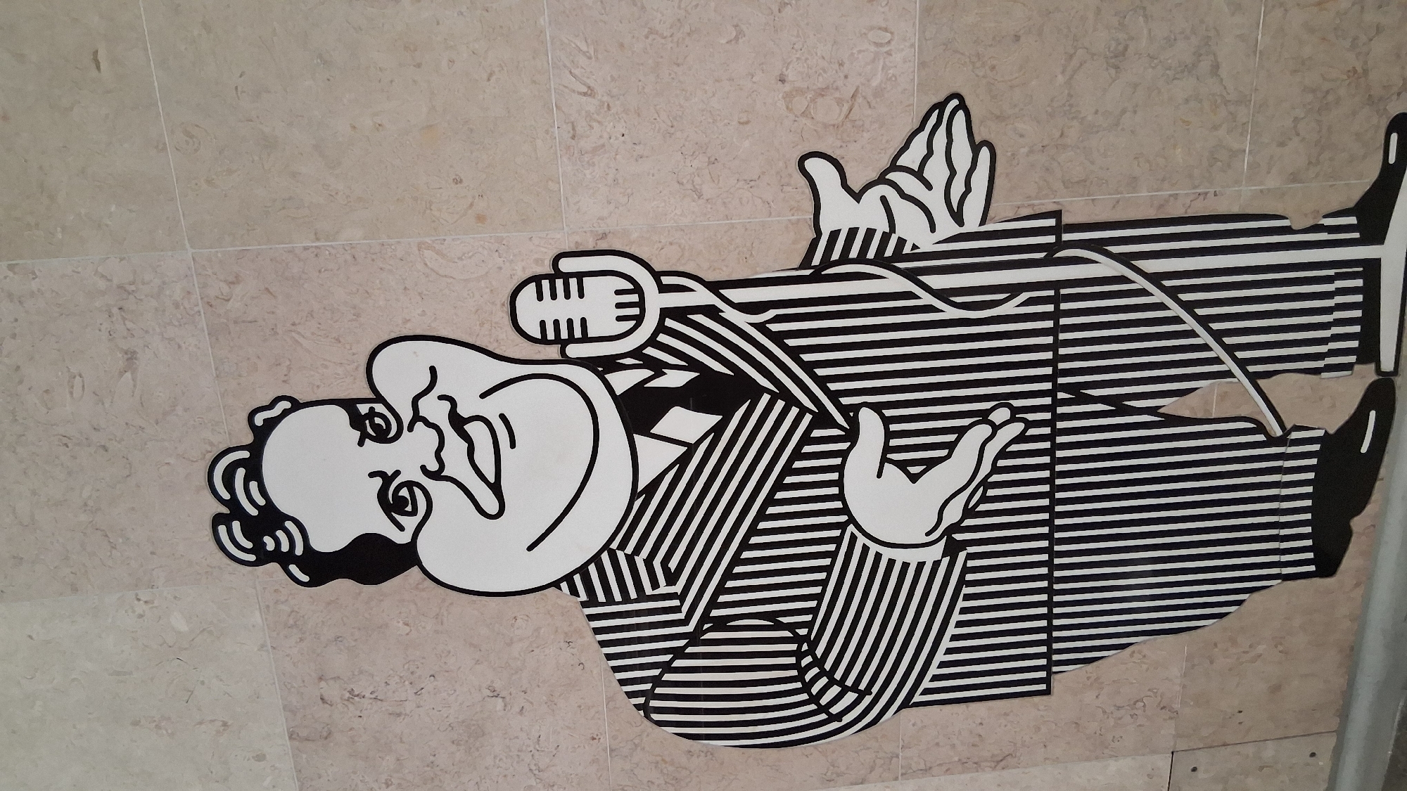 This image shows a large, stylized black and white graphic of a man in a pinstripe suit holding a microphone, positioned on a marble-tiled floor. The man has puffed-out cheeks, a wide smile, and curly hair. He is standing with his left hand extended and his right arm bent, holding the microphone. The graphic is placed on a light-colored marble floor with visible grout lines. This artwork is located in the Lisbon Oceanarium, in the Parque das Nações area of Lisbon, Portugal.