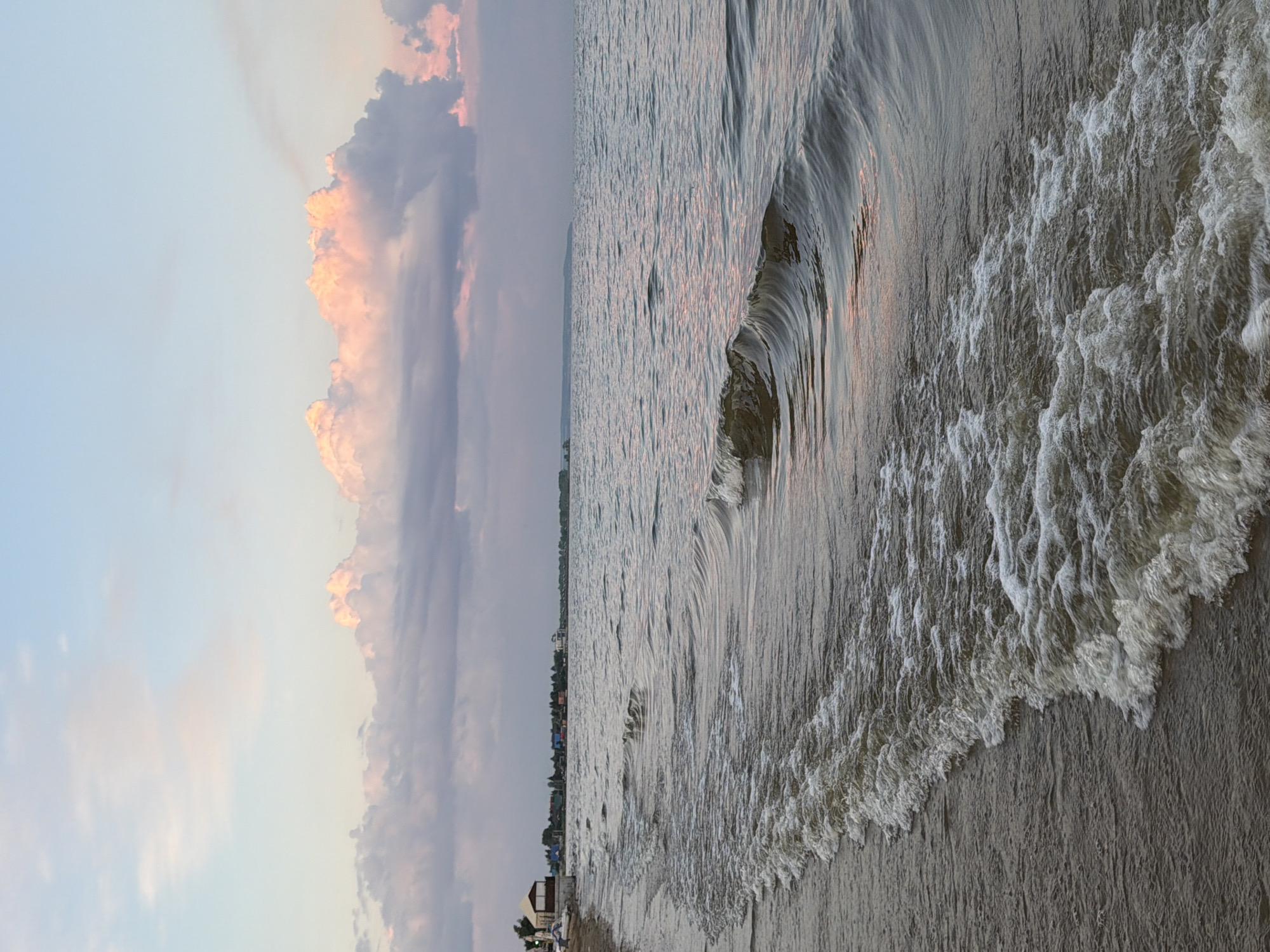 A beach scene in Laishevo, Russia. The image captures the shoreline of a body of water, likely the Kama River, with gentle waves lapping the sandy beach. In the distance, a line of trees and buildings suggests a settlement. The sky above is a soft blend of blue and pink hues, with fluffy clouds reflecting the warm light of either sunrise or sunset. The foreground is dominated by the textured surface of the water, showing the movement of the waves. There are no people or animals visible in the image.