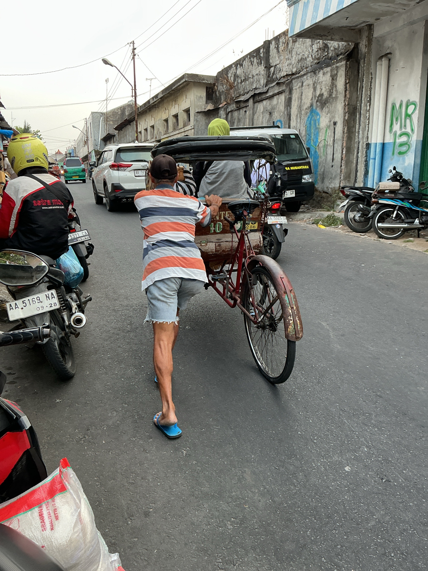 The image shows a street scene in Magelang, Indonesia. In the foreground, a man is pushing a pedicab. He is wearing a striped t-shirt and denim shorts, and blue flip-flops. The pedicab is red and appears to be old and somewhat rusty. Behind the pedicab, a white SUV is visible, along with several motorcycles. The street is paved and appears to be busy with traffic. Buildings line one side of the street, some with peeling paint and graffiti. The sky is overcast. The overall impression is of a typical urban environment in Indonesia.