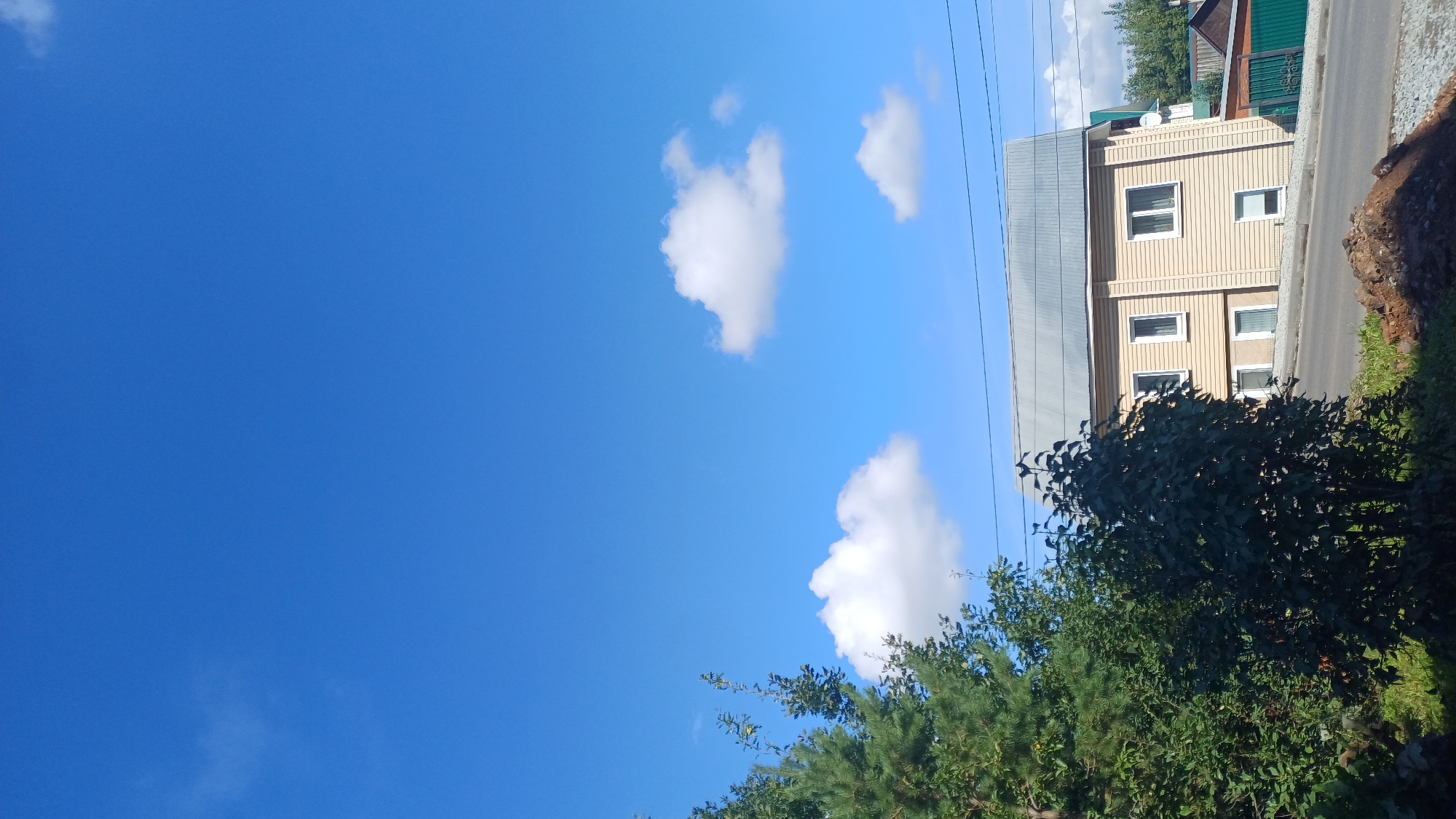 This is a vertical, eye-level outdoor shot featuring a two-story beige wooden house with a grey metal roof. The house has several white-framed windows, with two on the upper level and three on the lower level, two of which are partially obscured by the exterior siding. The house is situated on a paved road or driveway, with a patch of dark soil and gravel in the foreground. To the left of the house, there are dense green trees and foliage, including what appears to be a pine tree. In the background, there are more trees and glimpses of other buildings, including a green fence. The sky is a clear, vibrant blue with a few scattered white cumulus clouds. The overall scene suggests a residential area in a temperate climate, likely during daytime given the bright sunlight. The image does not contain any people or overt actions. Based on the architectural style and surrounding vegetation, this scene could be representative of a typical residential street in Izhevsk, Russia.