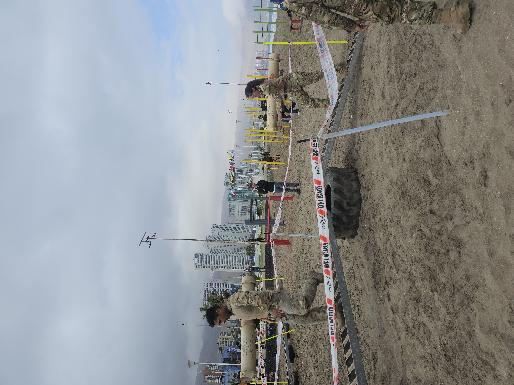 The image shows a sandy obstacle course with several individuals dressed in camouflage uniforms. In the foreground, two individuals are actively participating in an exercise, each carrying a large cylindrical object. A man on the left is walking, holding the cylinder horizontally across his shoulders. A woman on the right is also holding a similar cylinder, with her body angled as if she is in motion. Behind them, other people are visible, some standing and observing, while others appear to be setting up or managing the course.
The obstacle course features various elements, including a large tire, metal ramps, and yellow poles, all set up on the beach. Red and white caution tape, marked with "PELIGRO" (Danger in Spanish), is strung across parts of the course, indicating a restricted area or a hazard.
In the background, the cityscape of Iquique, Chile, is prominent, with numerous tall buildings lining the coast. The ocean is visible beyond the beach, and mountains form the backdrop. The sky is overcast, suggesting a cloudy day. The overall scene depicts a physical training or competition event taking place on a beach in Iquique.