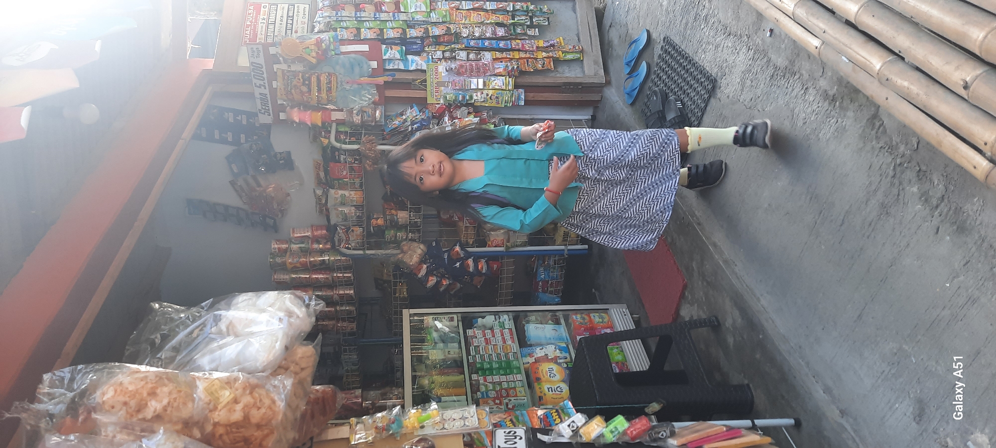 A young girl stands in front of a small storefront in Ploso, Indonesia. She is wearing a teal top and a patterned skirt, with yellow socks and dark shoes. Her hair is dark and falls around her face. The storefront appears to be a convenience store or small market, with shelves stocked with various goods, including packaged snacks, drinks, and household items. Large bags of snacks are visible hanging to the left of the girl. A black plastic stool is to her left. Outside the store, a red mat is on the ground, along with a pair of blue flip-flops and black sandals. Several bamboo poles are visible in the lower right portion of the image. The overall scene suggests a bright, sunny day.
