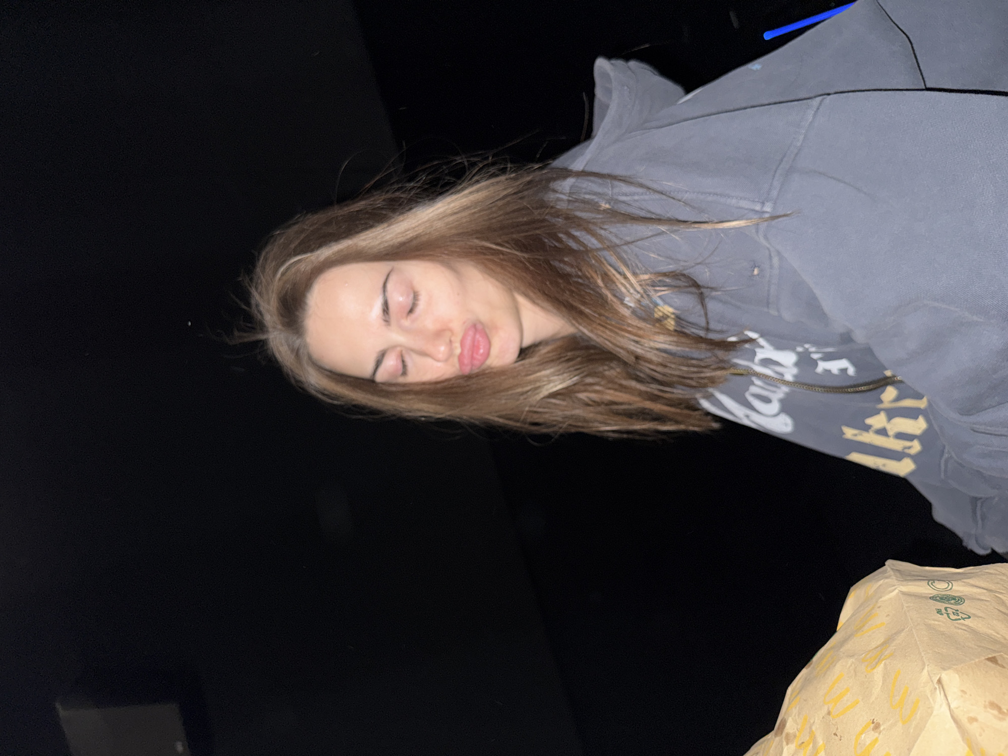 A person is depicted in a photograph, lying on their back with eyes closed and tongue slightly protruding. They are wearing a hooded sweatshirt with visible graphic elements on the chest. The background is primarily dark, with a partially visible paper bag in the lower right corner. The location is unknown.
