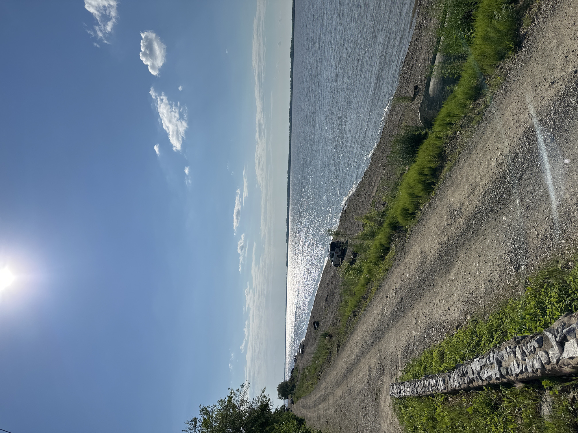 This is a photograph of a scenic view along the shore of a large body of water, likely a river or a lake, under a bright, sunny sky. The foreground features a dusty, unpaved road that curves to the left, bordered by a low, dry-stone wall on its left side. Green grass and some bushes grow along the roadside and on the embankment.
The road leads towards the water, which occupies the right side of the frame. The water is a deep blue and reflects the bright sunlight, creating a shimmering, glittering effect on its surface. The shoreline is a mix of pebbles and sand, with some sparse green vegetation.
In the middle ground, parked on the shoreline, are several vehicles: a dark SUV, and a lighter-colored SUV. There are also some scattered objects on the beach, including what appears to be a tire. Further in the distance, along the horizon, a faint line of trees and structures can be seen. The sky is a clear, vibrant blue with a few scattered white clouds. The sun is high in the sky and is very bright, causing a lens flare in the lower right portion of the image.
The scene depicts a natural, somewhat remote location, suggesting it could be a recreational area or a less developed coastal region. The presence of vehicles indicates human activity, possibly for fishing, camping, or simply enjoying the outdoors. There are no people clearly visible in the image. The overall impression is one of a peaceful, sun-drenched day by the water.