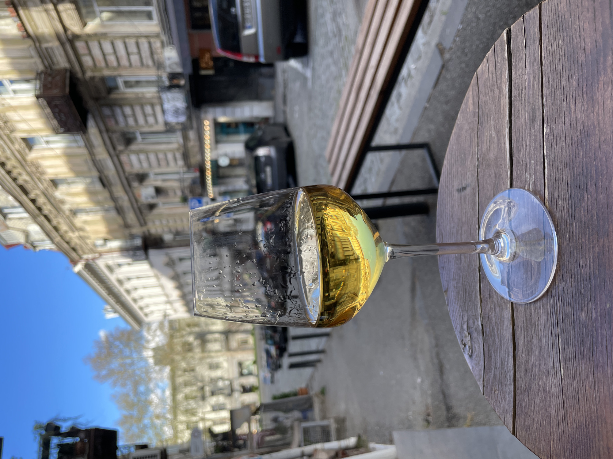 A wine glass filled with a light-colored liquid rests on a round, wooden tabletop outside in Tbilisi, Georgia. The background shows European-style buildings and a street with parked cars and pedestrian traffic. The weather appears sunny and mild.
