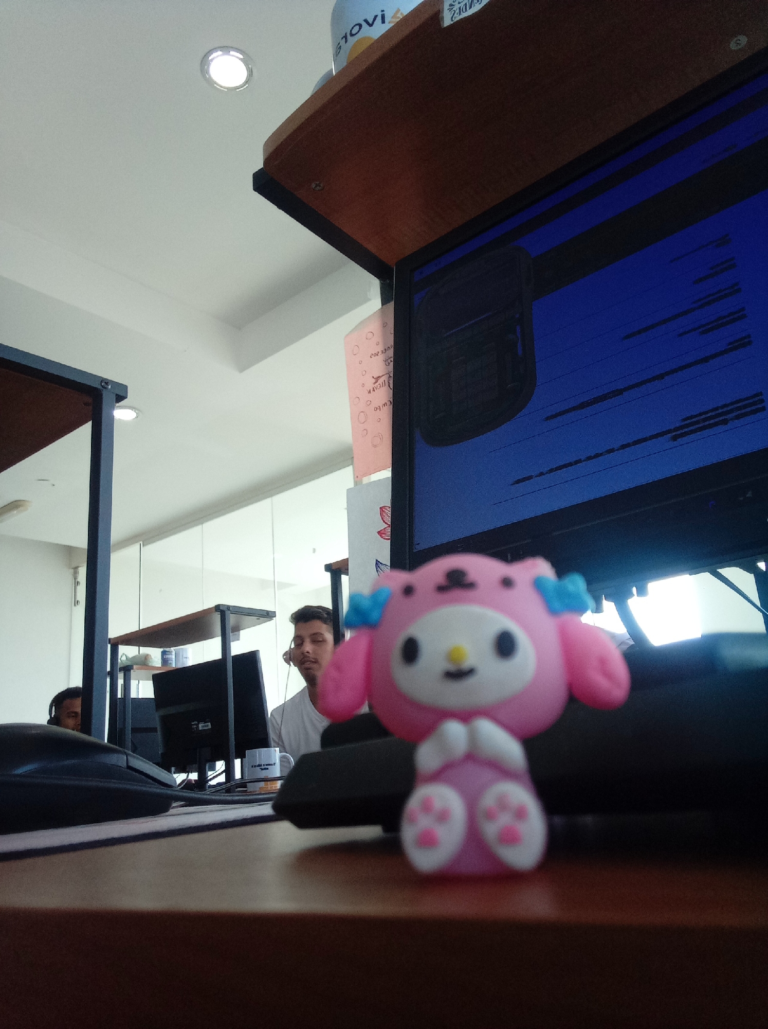 A dimly lit office in Parroquia Guatire, Venezuela, features a desk with a pink My Melody figurine in the foreground. A computer monitor, keyboard, and mouse are also on the desk. Two men are visible in the background, wearing headsets and working at computer stations. A shelf with a bottle is positioned above the monitor. The room appears to be a workspace with multiple desks.