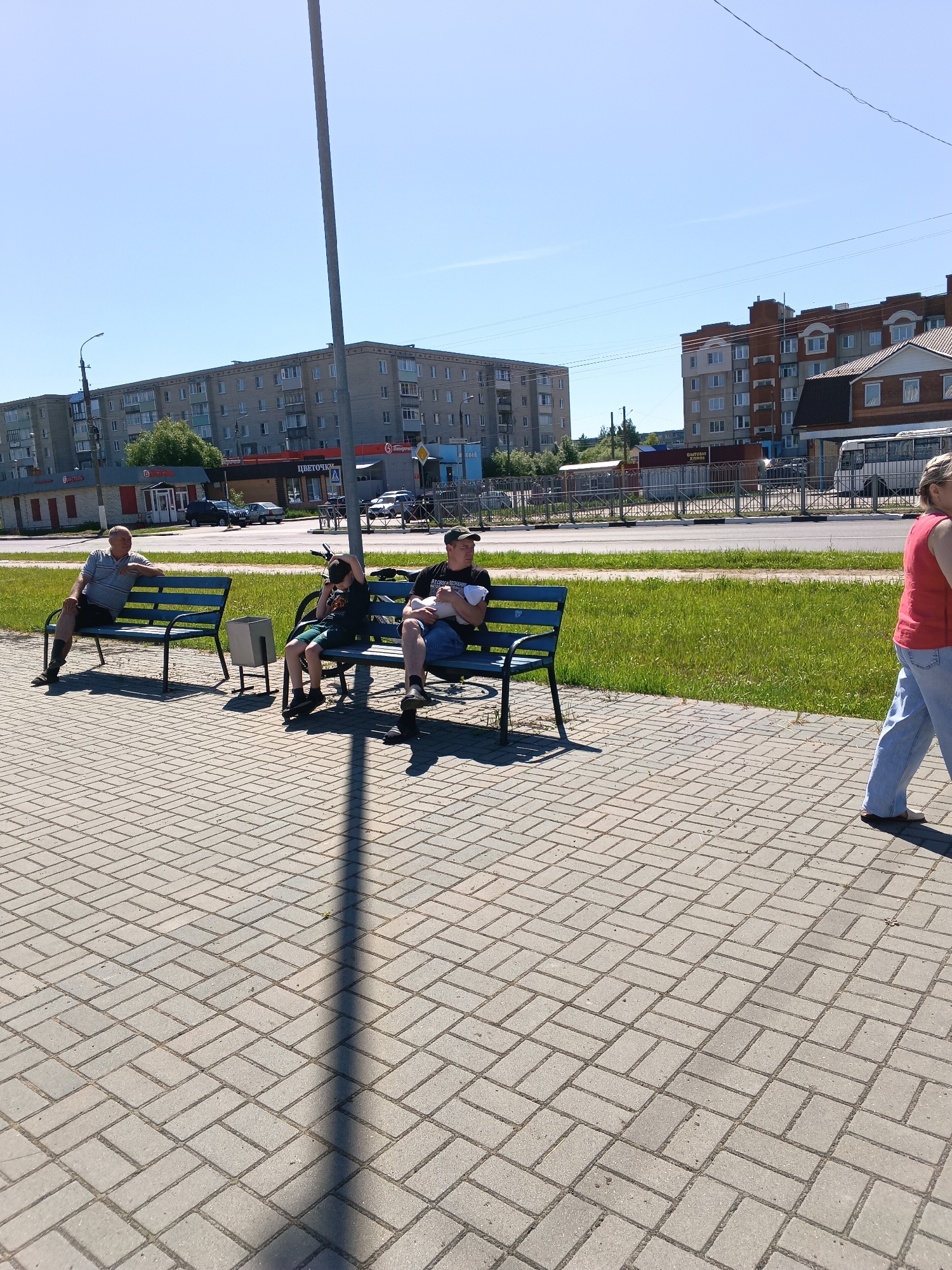 The image depicts an outdoor urban scene in Sudogda, Russia, on a bright, clear day, likely midday or early afternoon given the strong shadows. The foreground features a light grey brick-patterned paved pathway, laid in a herringbone design, with a prominent grey lamppost casting a long, diagonal shadow to the lower right.
Three individuals are seated on two blue metal benches with wooden slats. On the left bench, an older man with grey hair, wearing a light-colored striped short-sleeved shirt, dark shorts, and dark socks, sits relaxed, looking towards the right. Between the benches, a rectangular grey metal garbage bin is visible. On the right bench, a young boy in a dark t-shirt and green shorts rests his head on his hand, looking downwards; a bicycle is leaning against the back of this bench. Next to him, an adult man wearing a dark t-shirt and a dark cap is holding a baby wrapped in a white blanket. He is looking towards the right, away from the camera. A woman, partially visible on the far right, walks from right to left, facing away, wearing a red sleeveless top, light blue jeans, and dark flat shoes.
Behind the paved area and benches, a wide strip of vibrant green grass separates the pathway from an asphalt road. A metal guardrail or fence runs parallel to the road. In the background, multiple multi-story residential buildings with grey and reddish-brown facades are visible. Several commercial storefronts line the ground floors of these buildings, displaying signs in Cyrillic, including "Пятерочка" (Pyaterochka, a supermarket chain), "ЦВЕТОЧКИ" (Tsvetochki, meaning Flowers), and "БЫТОВАЯ ХИМИЯ" (Bytovaya Khimiya, meaning Household Chemicals). Various vehicles, including cars and a white bus, are present on or beside the road. The sky is a clear, cloudless blue.