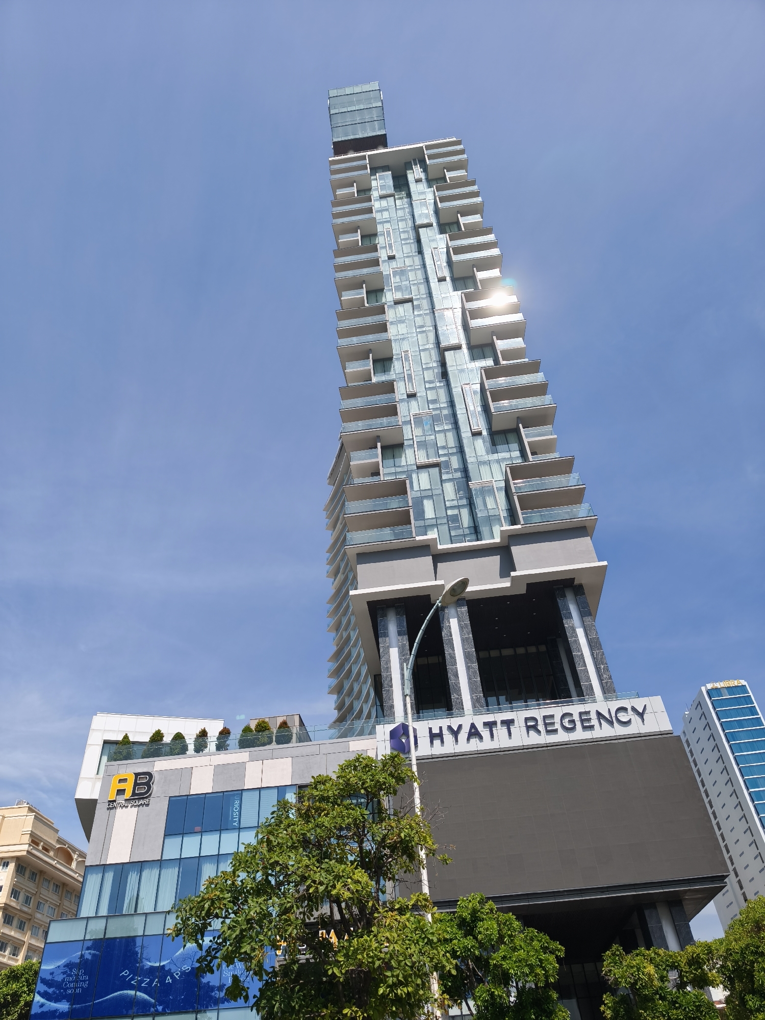 The image captures a low-angle view of the Hyatt Regency hotel in Nha Trang, Vietnam. The modern, glass-faced hotel building dominates the frame, with its balconies and sleek design reaching towards a clear blue sky. Sunlight glints off the glass facades, creating a bright, sun-drenched atmosphere. To the left of the Hyatt Regency, the "AB Central Square" building is visible, featuring a prominent "AB" logo and large glass windows. Lush green trees partially obscure the lower sections of the buildings, adding a touch of nature to the urban landscape. In the background, other buildings contribute to the city's skyline. The scene appears to be a daytime shot on a clear, sunny day.