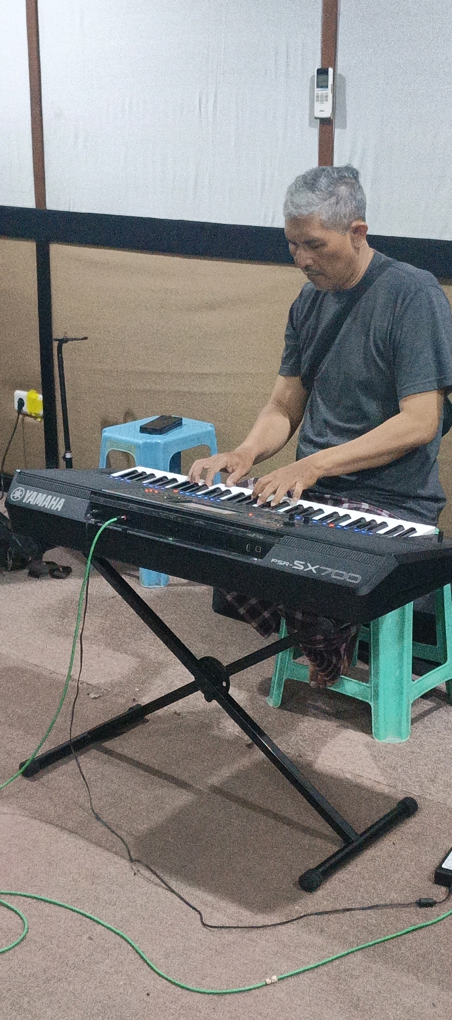 An older man with graying hair is playing a Yamaha PSR-SX700 keyboard. He is seated on a turquoise plastic stool in what appears to be a makeshift studio or practice space. The background features a plain, beige wall and a white screen or backdrop. A microphone stand is visible to the left, and an air conditioning remote is mounted on a wooden beam above the man's head. The keyboard is supported by a black, X-shaped stand, with a green cable running from the keyboard to the floor. The floor is carpeted in a light brown color. The man is wearing a dark gray t-shirt and plaid pants. The scene is likely located indoors, possibly in a residential area or small venue in Surabaya, Indonesia, given the typical furnishings and overall atmosphere.