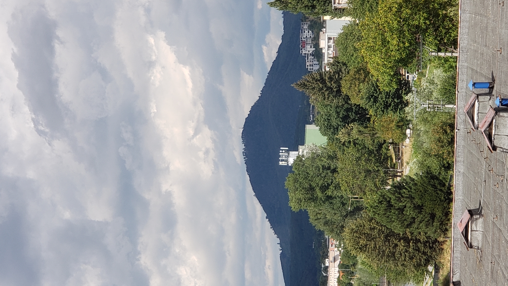 The image captures a panoramic view of a dark, tree-covered mountain against a cloudy sky. At the peak of the mountain, several small structures, possibly antennas or small buildings, are visible. In the mid-ground, a white tower with multiple antennas stands prominently in front of a light green building. Behind this, several modern white buildings with balconies form a residential area. The foreground shows a densely wooded area with a variety of trees, and a glimpse of a body of water to the left. The very front of the image reveals the flat, grey, weathered roof of a building with several small dormer-like structures and a bright blue chimney. The scene appears to be a landscape view from a city, with the mountain dominating the background. This landscape strongly resembles the view of the Šariš Castle hill, a prominent landmark located just outside the city of Prešov, Slovakia.
