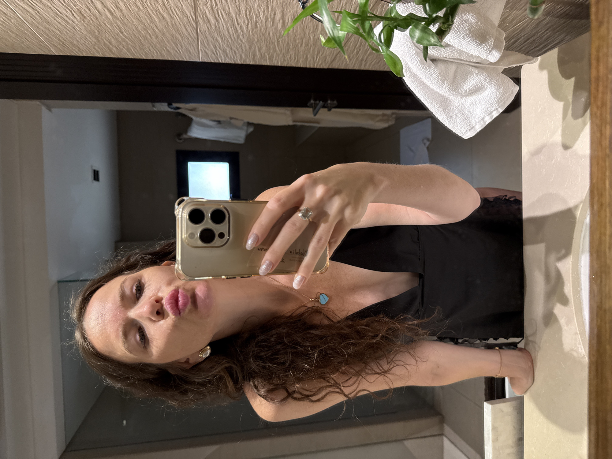 A woman is taking a selfie in a bathroom mirror. She has long, curly brown hair and is wearing a black slip dress, a blue heart-shaped necklace, and earrings. She is puckering her lips for the photo and holding her phone, which has a gold case, in her right hand. Her fingernails are painted with a glittery polish. A ring is visible on her left hand. The bathroom appears to be luxurious, with tiled walls and a modern sink. There is a folded white towel on the right side of the sink, and a plant with green leaves is visible. In the background, there are bathrobes hanging on a rack and a television screen. The scene suggests the woman is either at home or in a hotel room in Dubai, United Arab Emirates.