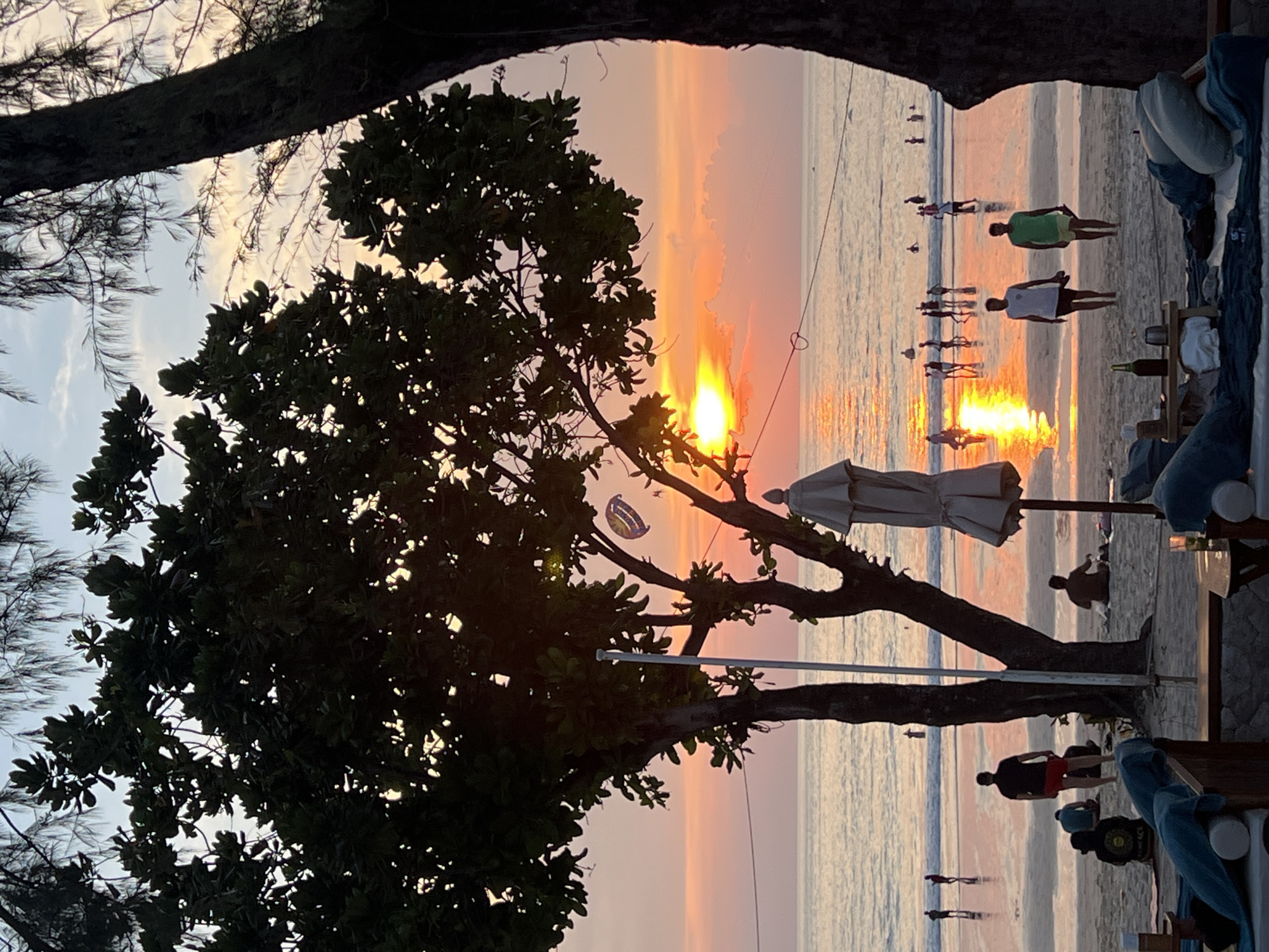 In Patong, Thailand, a vibrant sunset illuminates a tranquil beach scene. The sky, a canvas of warm oranges and soft pinks, reflects on the gentle waves of the Andaman Sea. Silhouetted against this breathtaking backdrop, people engage in various activities: some stroll along the shoreline, others wade in the water, and a few relax on beach chairs in the foreground. Lush green trees frame the view, their leaves rustling in the evening breeze. A parasail floats lazily in the distance, adding a touch of adventure to the serene atmosphere. The overall impression is one of peaceful enjoyment, a perfect end to a day in paradise.
