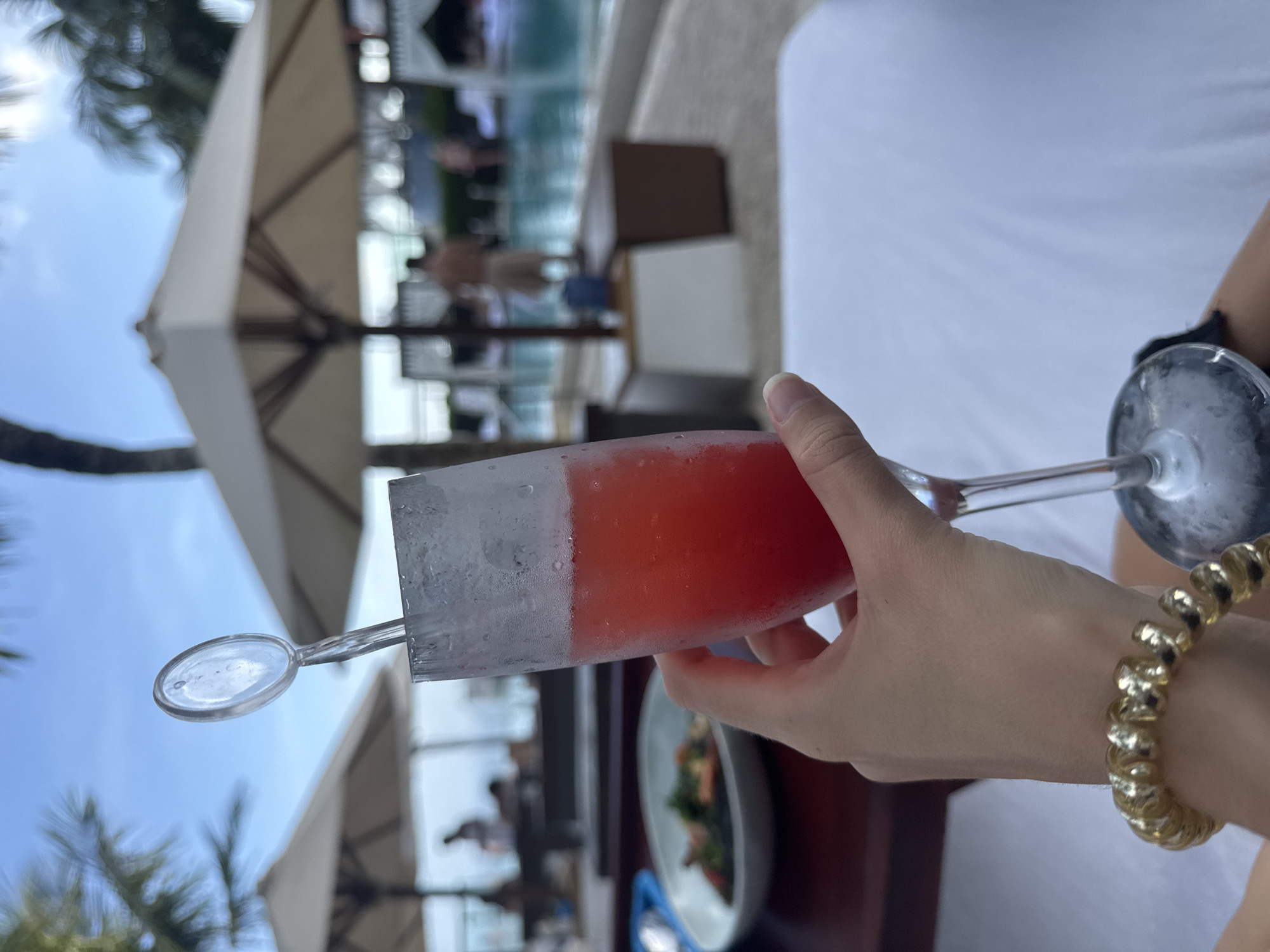 The image shows a person holding a chilled glass of red liquid, likely a cocktail, by a poolside. The location is suggested by the presence of palm trees, sun umbrellas, and a swimming pool in the background, potentially in a tropical or coastal resort. The person is wearing a gold bracelet on their wrist. A plate with food is visible on a table nearby. The image is shot at a slightly rotated angle.