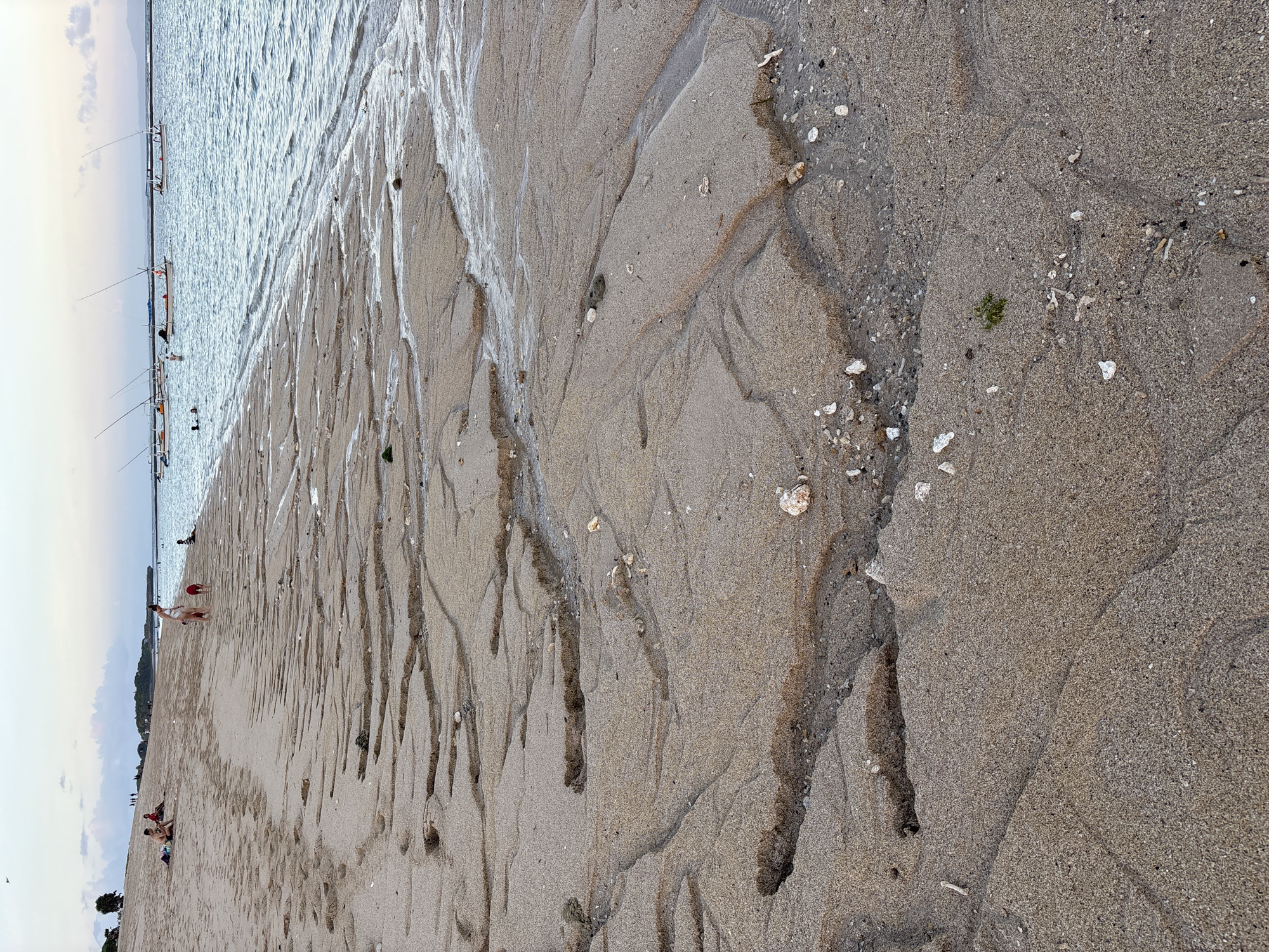 This horizontal image depicts a beach scene in Kuta Selatan, Indonesia. The foreground is dominated by a sandy beach with distinctive water erosion patterns, forming rivulets and ridges. Scattered small shells and debris are visible on the sand. In the background, the ocean stretches towards the horizon. Several small boats are anchored in the shallow water near the shore. People are seen both on the beach and in the water, with some standing near the boats. The sky is overcast, casting a soft, diffused light over the scene.