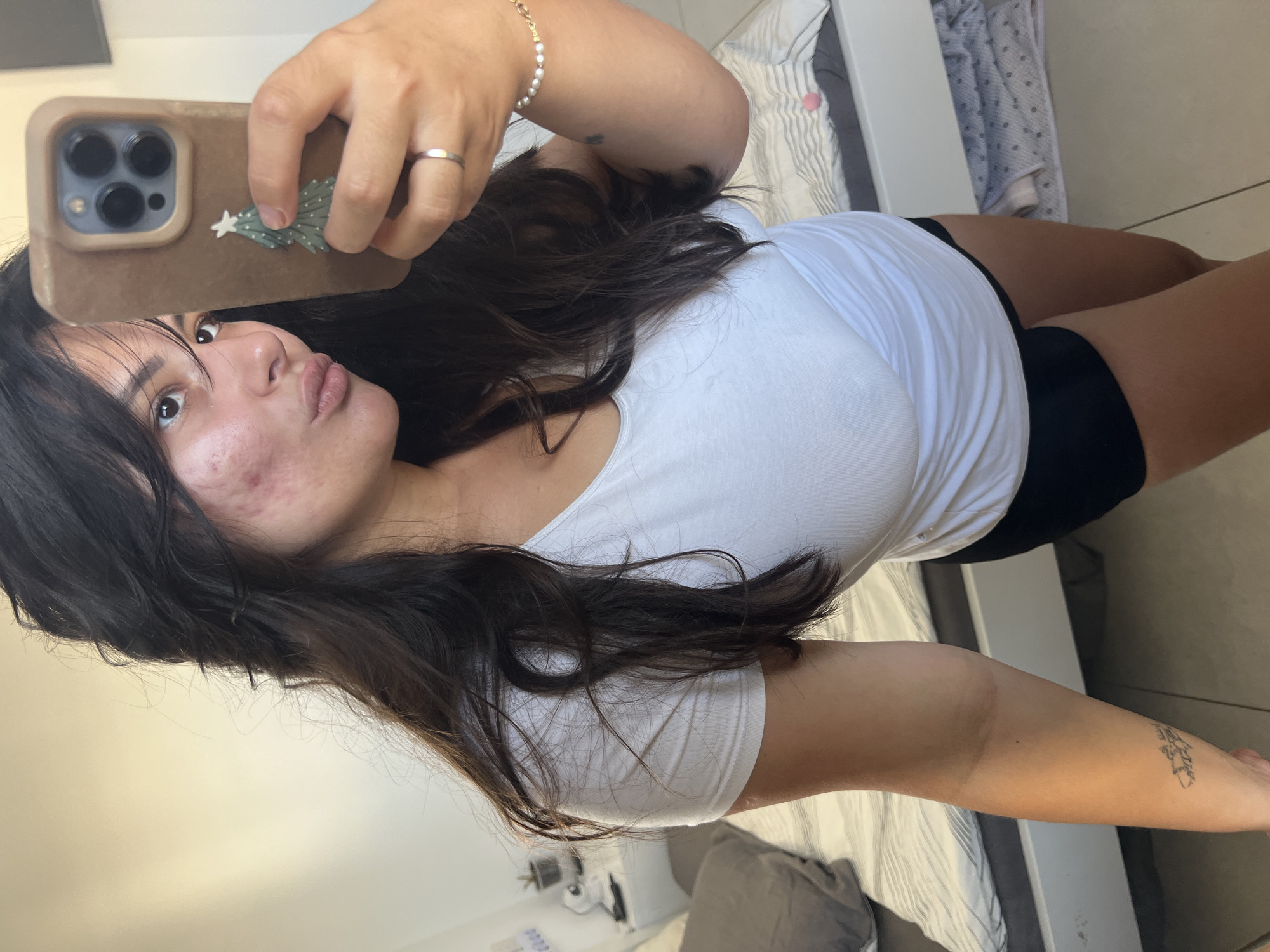 A young woman with long, dark hair is taking a selfie in a bedroom. She is wearing a white t-shirt and black shorts. She has acne on her face. She is holding a smartphone with a case featuring a Christmas tree design. There is a ring on her left ring finger and a bracelet on her right wrist. The room appears to be a bedroom with a bed in the background. The overall setting suggests it is a personal space. The image was taken in Kiryat Bialik, Israel.