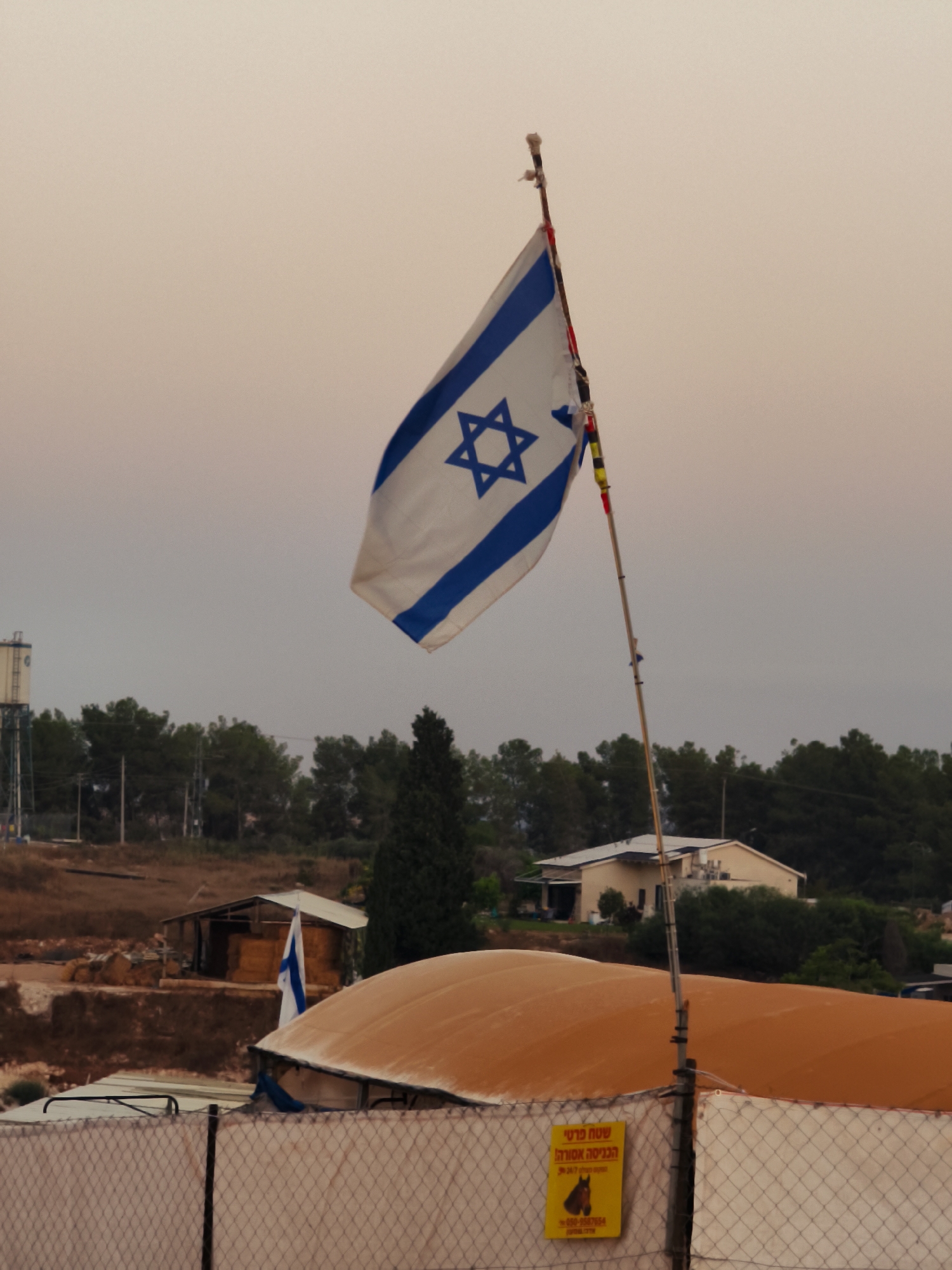 An Israeli flag flies from a tall pole in front of a dusky sky. In the background, a landscape features a water tower, trees, and a house with a light-colored roof. A fence with a diamond pattern runs across the foreground. A yellow sign with Hebrew text is attached to the fence. The scene is likely located in the Lachish Regional Council, Israel.
