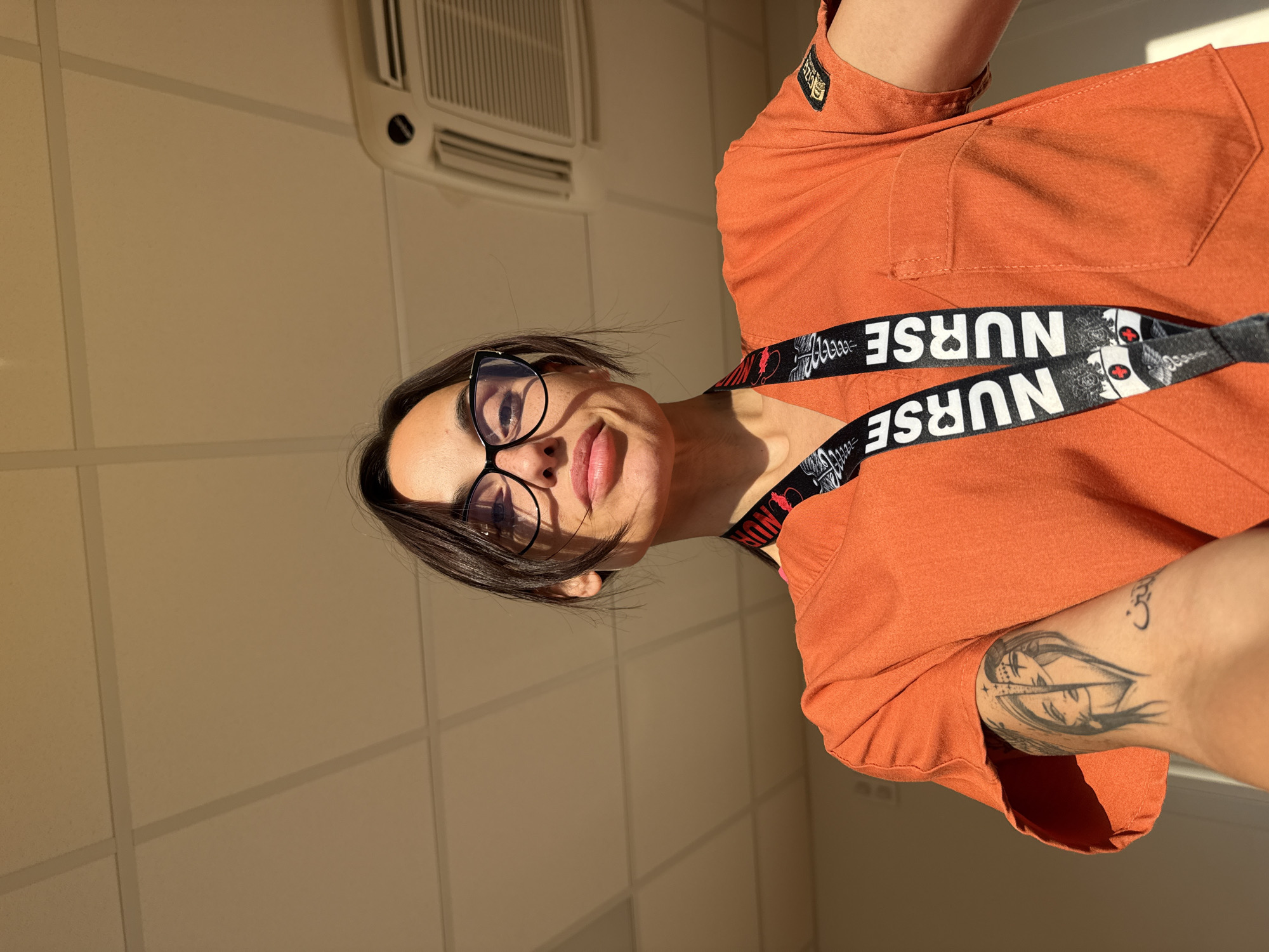 A woman wearing an orange scrub top and black-rimmed glasses is taking a selfie. She has dark hair pulled back and is looking directly at the camera with a slight smile. A lanyard with the word "NURSE" repeated on it is visible around her neck. She has a tattoo on her left arm. The background appears to be indoors, with a white tiled ceiling and an air conditioning unit visible. The lighting suggests it is daytime, possibly with sunlight coming in from a window. The location is not identifiable as Pleven, Bulgaria.