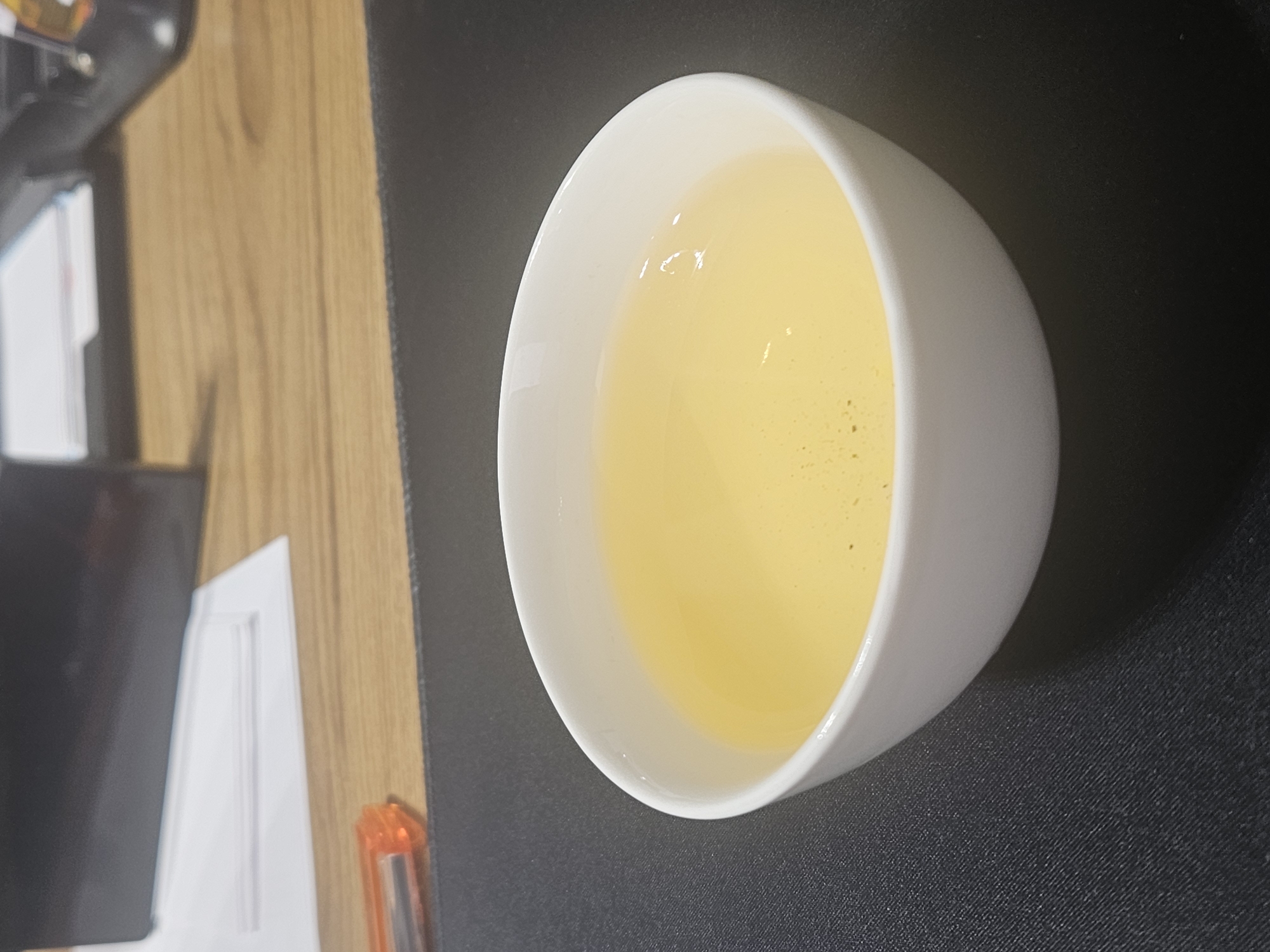 The image shows a white ceramic cup filled with a pale yellow liquid, likely tea, on a dark gray textured surface. The cup is positioned slightly off-center, towards the bottom right of the frame. In the background, a wooden surface is visible, with a stack of papers and a portion of what appears to be a black electronic device. An orange pen or marker is also partially visible on the wooden surface. The overall scene suggests an indoor setting, possibly an office or a desk area. There are no people or animals present in the image. The lighting suggests it is daytime, with some reflections visible on the surface of the liquid in the cup. The location in the city of Uchtepa, Uzbekistan, is not discernible from the visual information provided in the image.