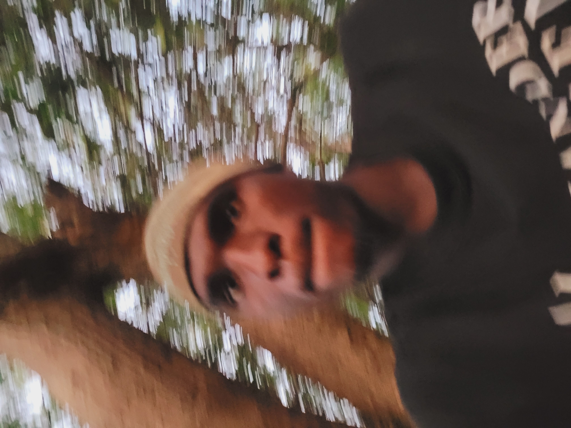 The image is a blurry, low-resolution selfie taken outdoors. A person with dark skin and wearing a light-colored head covering is in the foreground, looking towards the camera with their mouth slightly open. They are wearing a dark t-shirt with partially visible white lettering on the lower right side. To the left of the person, a large, textured tree trunk is visible. The background consists of blurred green foliage and bright white sky, suggesting the image was taken in a natural setting, possibly a park or wooded area. The motion blur indicates movement during the capture of the photo. There are no identifiable landmarks that place the image specifically in Bukuru, Nigeria.