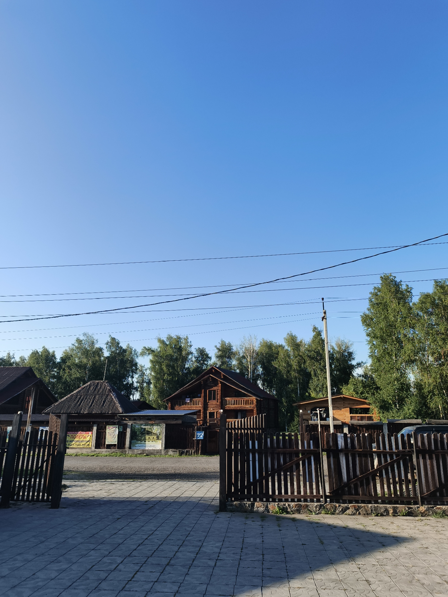 The image shows an outdoor scene with a clear blue sky and several wooden buildings. In the foreground, there is a paved walkway and a wooden fence. Behind the fence, there are various wooden structures, some appearing to be houses or cabins, with trees in the background. Several power lines stretch across the sky. There are also some signs visible on the buildings. The overall impression is of a rural or semi-rural setting, possibly a resort or a village, bathed in sunlight.