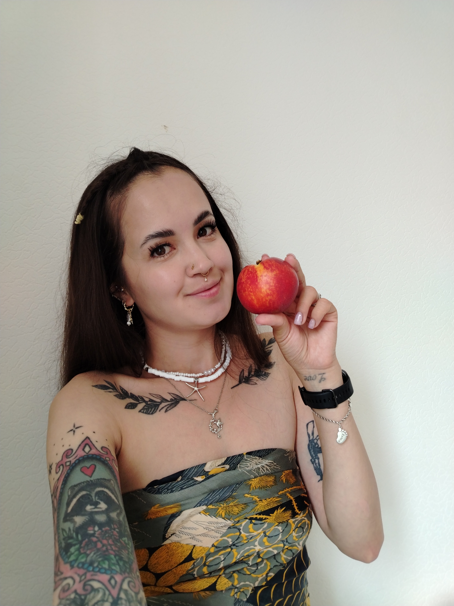 A young woman with long, dark hair and tattoos on her arms and chest holds a nectarine up to her face with her right hand. She is wearing a strapless top with a pattern of birds and foliage. She has on multiple necklaces, including a white beaded one and one with a starfish pendant. She also has earrings and nose piercings. The background is a plain, textured, off-white wall. The image appears to be a selfie taken indoors. There is no indication of the city of Noviy, Russia, or any specific location within it.