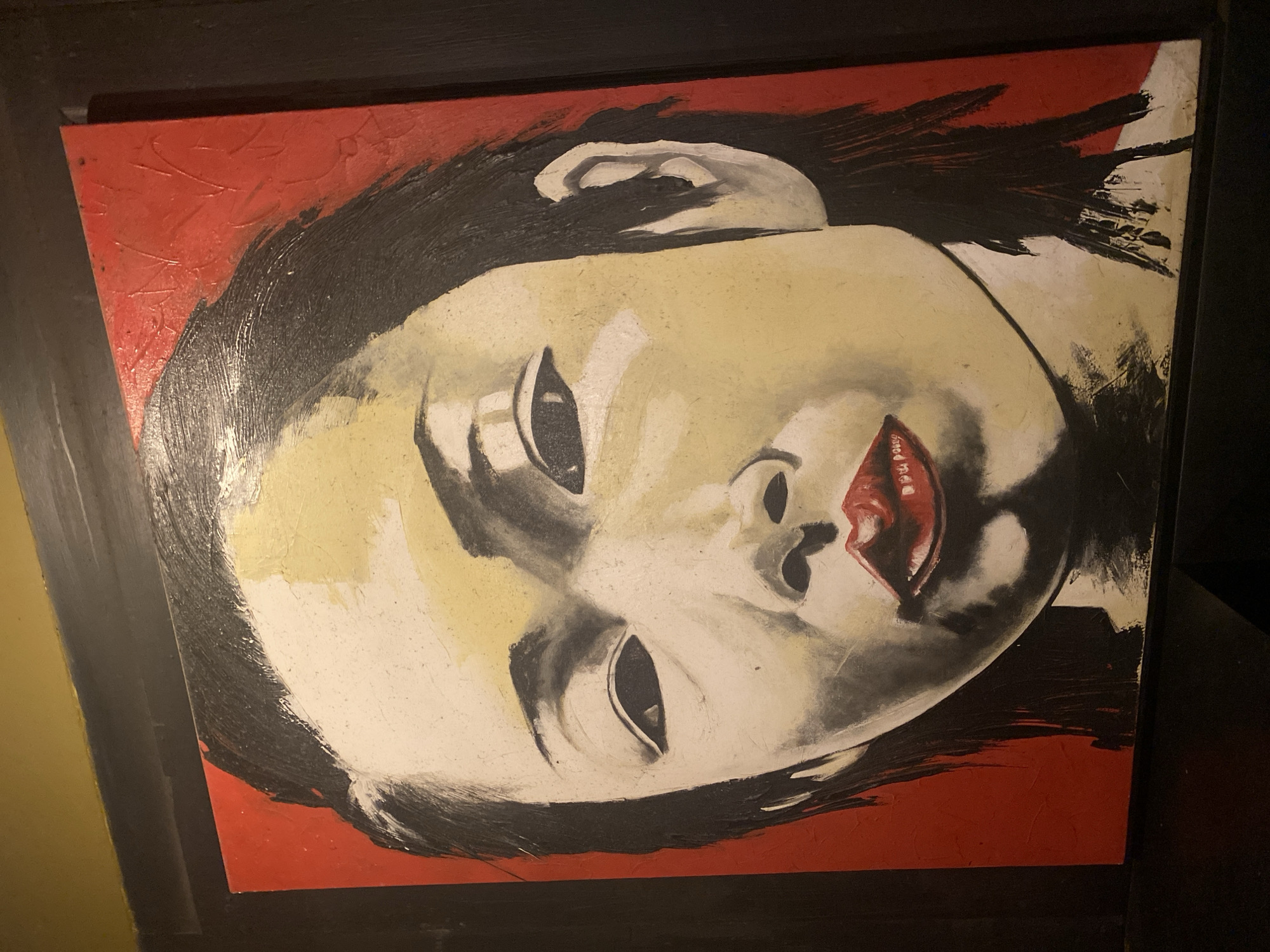 The image shows a close-up of a painting of a woman's face. The painting is in a realistic style, with bold brushstrokes and a limited color palette of red, yellow, black, and white. The woman has dark hair, dark eyes, and red lips. She is looking directly at the viewer. The background is a solid red color. The painting is hanging on a dark wall. It is unclear if this painting is located in the city of Denpasar, Indonesia, as there are no specific landmarks or contextual clues to suggest this location.