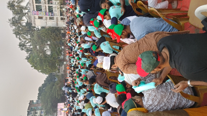 This image captures a large outdoor political rally or public meeting held in an urban space within Dhaka, Bangladesh. Thousands of adult men are densely packed, seated on individual light brown plastic chairs, facing towards an unseen stage or presentation area. A significant portion of the attendees wear identical light blue t-shirts, many featuring a logo on the back. Headwear varies, with many men wearing white or colored prayer caps (topi), and a notable number sporting red and green baseball caps.
In the mid-ground, a large screen displays Bengali text, including "নির্বাচনী জনসভা" (Election Public Meeting), indicating the event's nature. The background features multi-story concrete buildings, some appearing residential with clothes hanging from balconies. One prominent building displays a large, vertically oriented banner with colorful Bengali script. Various large trees, including palm trees, are interspersed among the buildings and rise above the crowd. The sky is hazy and light-colored. In the immediate foreground, a man in a black polo shirt and a red and green cap is visible from behind, while another man seated in front of him holds a light blue piece of fabric or a mask. The gathering appears orderly and well-attended.