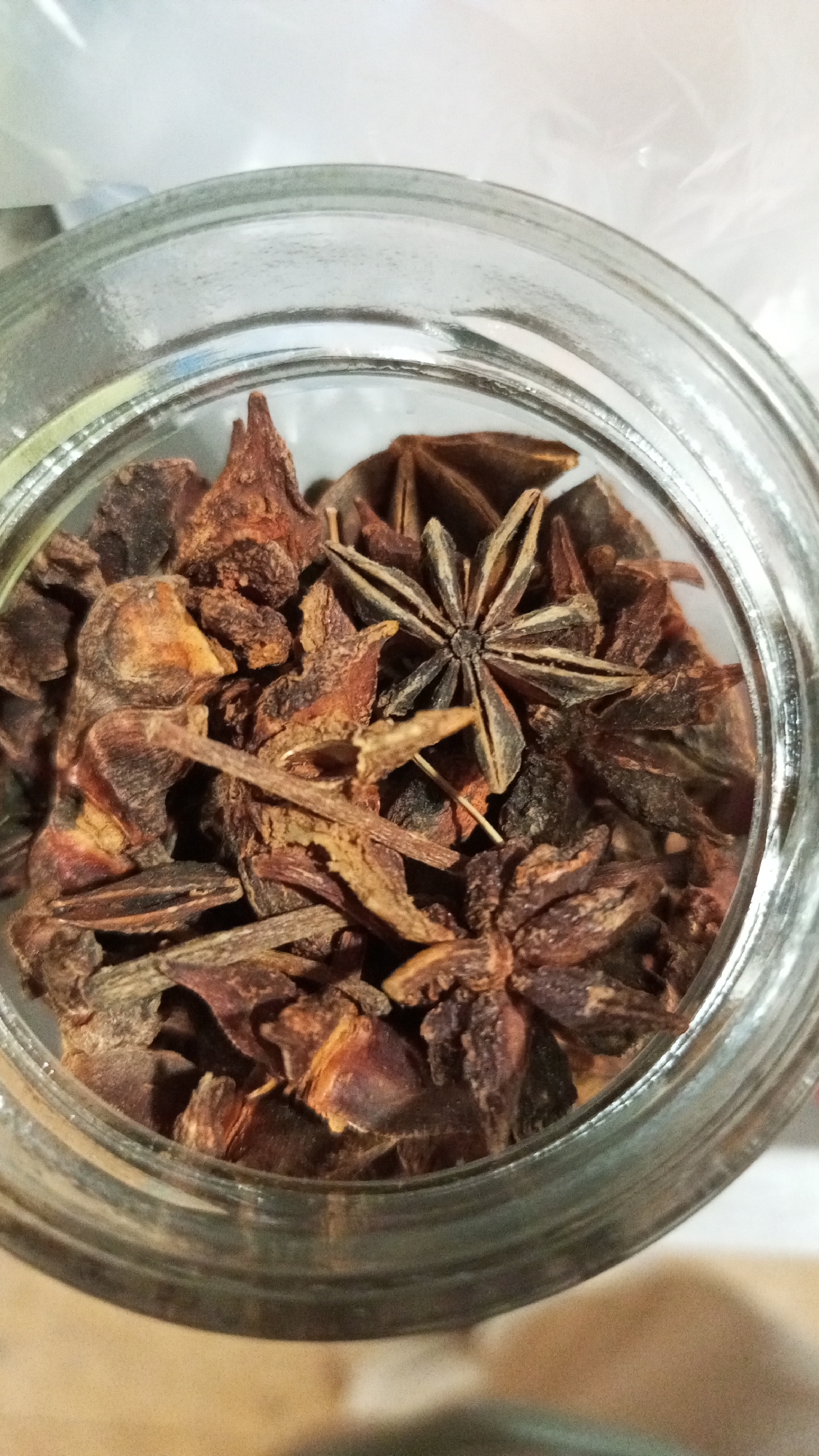 An overhead shot shows a clear glass jar filled with star anise and small pieces of bark. The star anise are mostly dark brown, with one lighter-colored star anise in the center. The bark pieces are also dark brown with some reddish hues. The background is blurred. The location cannot be determined from the image.