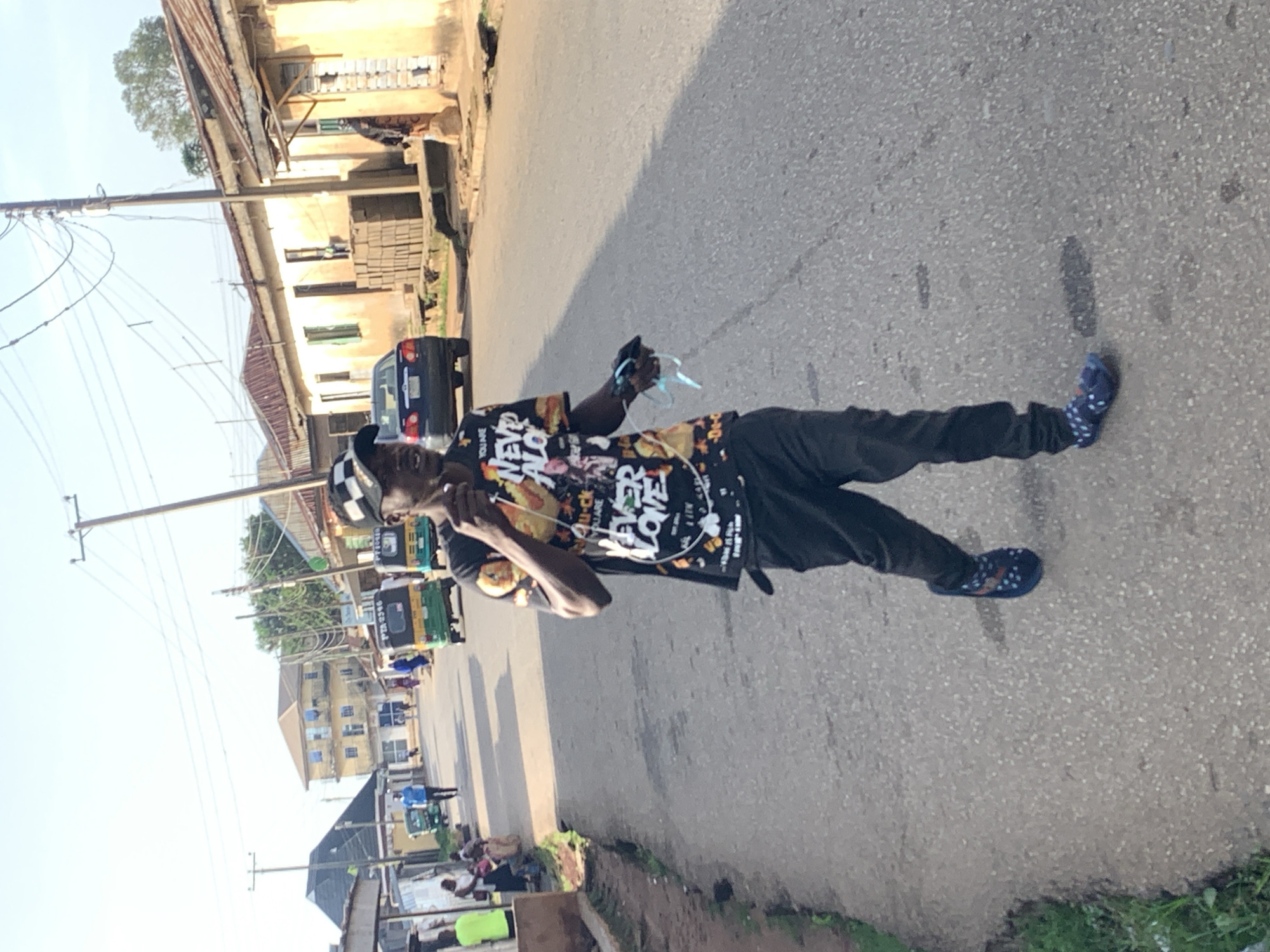In a street scene in Jos, Nigeria, a Black male individual walks forward on a paved road. He is wearing a black cap, a black t-shirt with a graphic design, black pants, and blue patterned slides. He holds a phone and what appears to be earbuds in his right hand. The road is lined with buildings, some with corrugated metal roofs, and there are several small motorized tricycles, known as Keke Napep, present. In the background, a dark-colored sedan is parked. People are visible in the background, some sitting on the side of the road and others walking. The sky is clear and bright.
