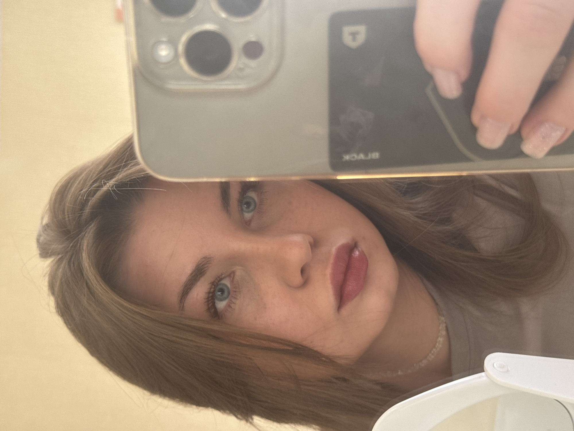 A young woman with light brown hair and blue eyes takes a selfie. She is holding a smartphone with a multi-lens camera and a black card case attached to the back. The card case has a logo resembling a 'T' in a shield and the word 'BLACK' printed in reverse. Her nails are light pink with glitter. A small, delicate necklace is visible on her neck. A white object appears in the lower right corner. The image provides no information related to the city of Chaikovsky, Russia.